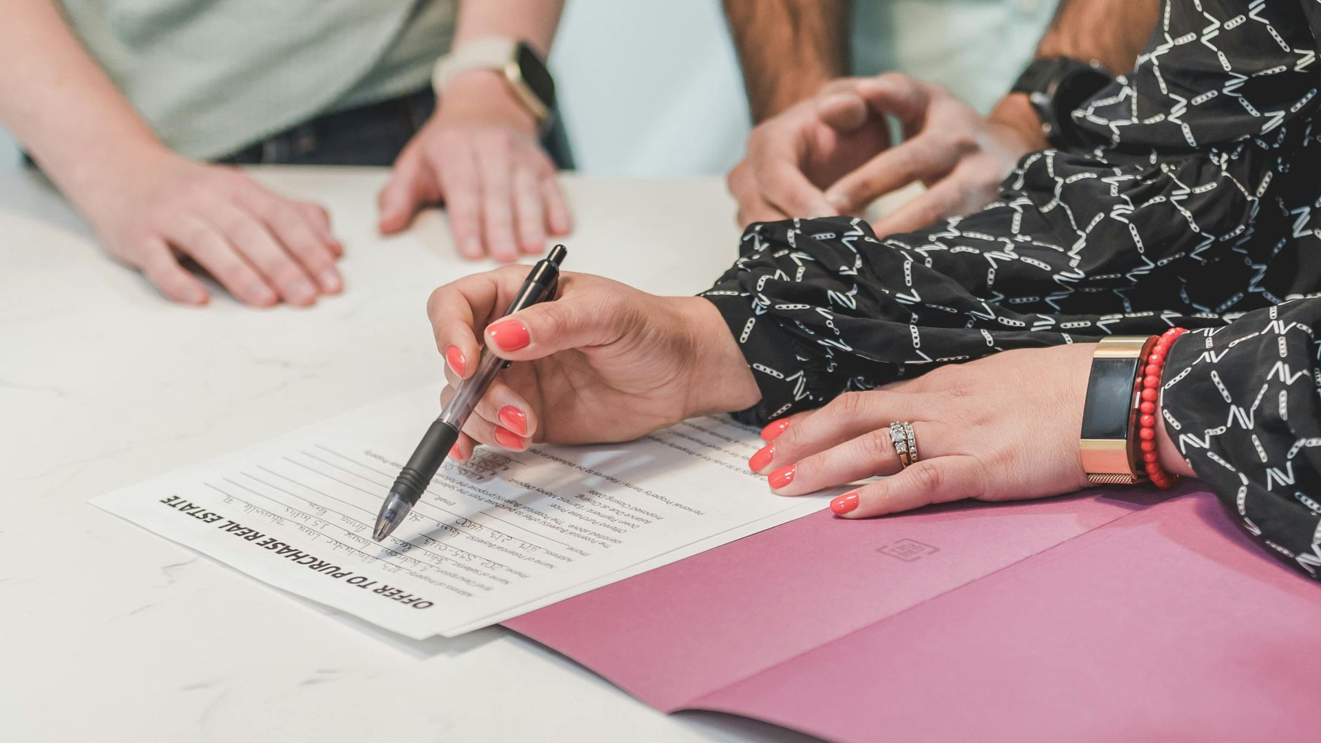 Close-up of hands signing a real estate document at a meeting table with three people.