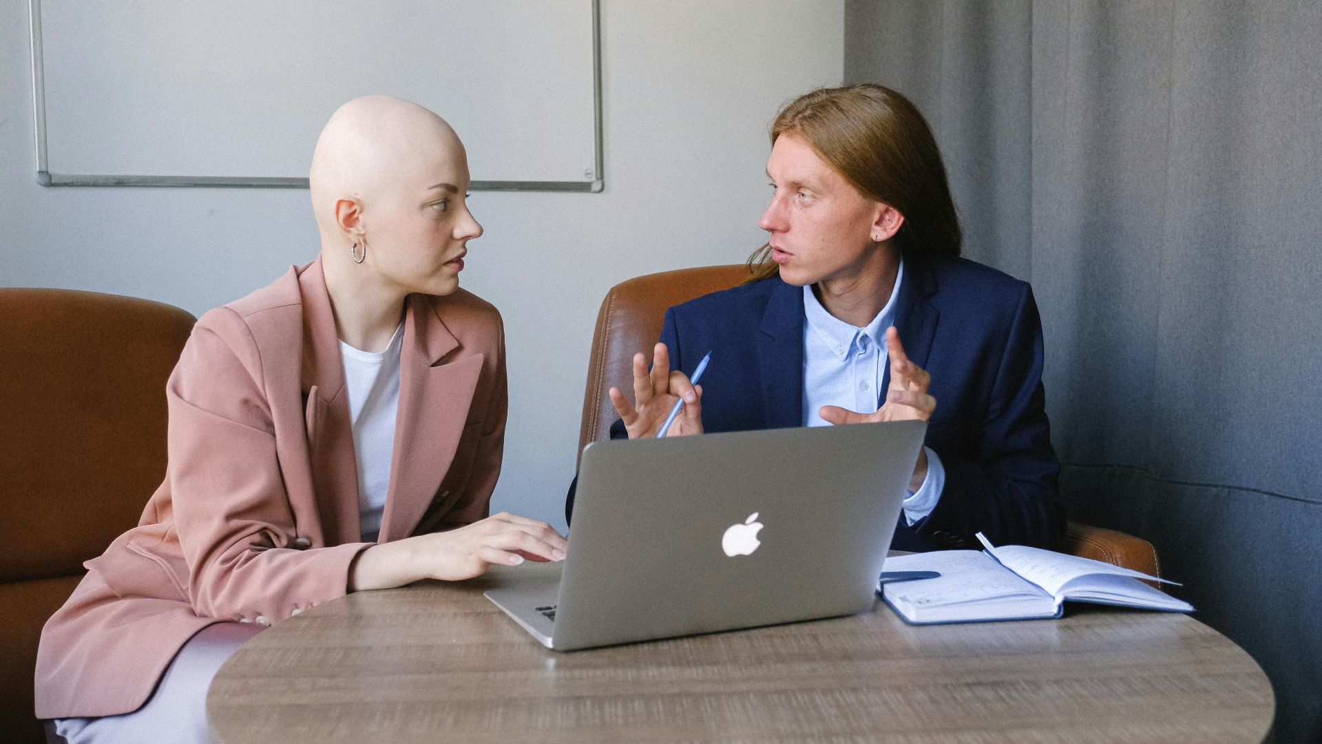 Concentrated young male employee sitting at round table with copybook and laptop while explaining to female colleague details of new project