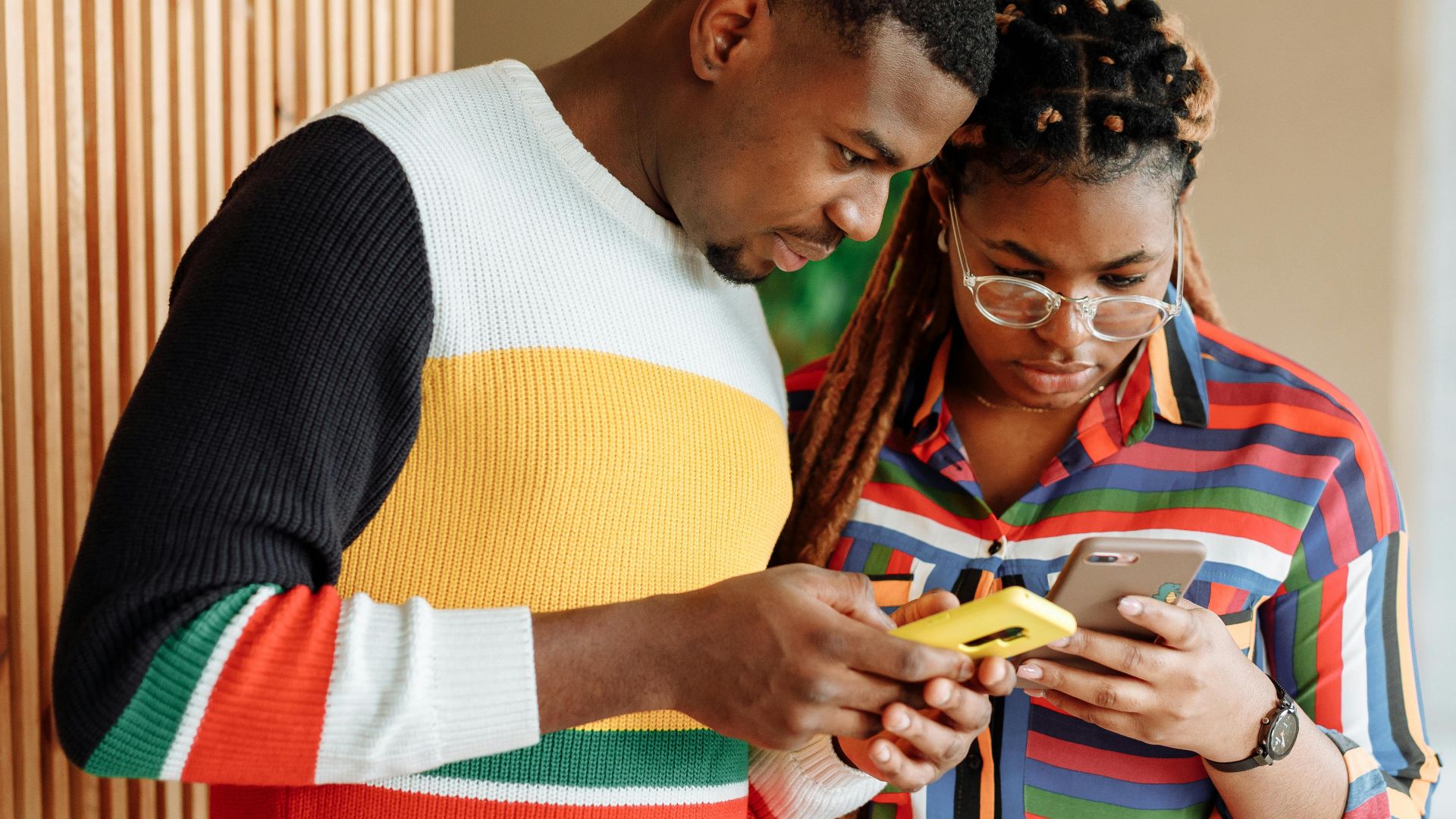 A young couple stands together indoors, focused on their smartphones, demonstrating connectivity and modern communication.