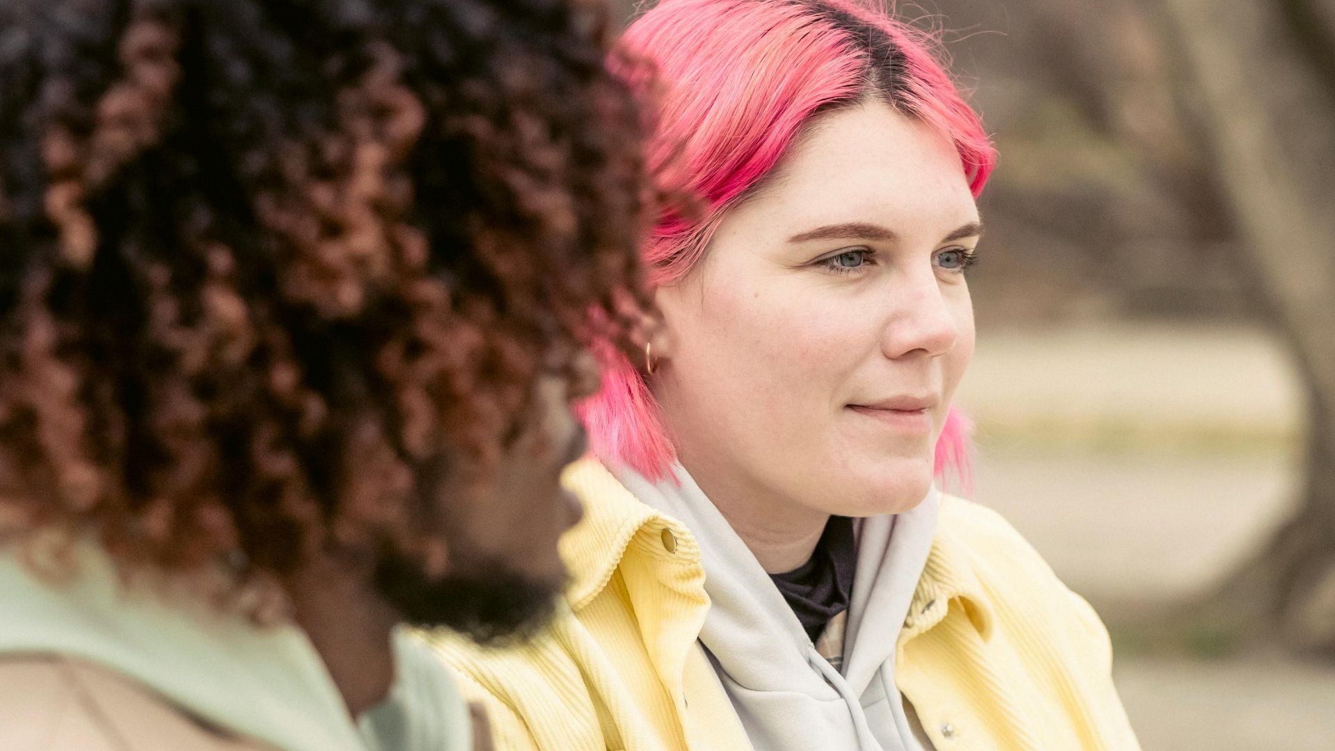 Calm woman with pink hair and anonymous African American man sitting on wooden table against building on street with tree