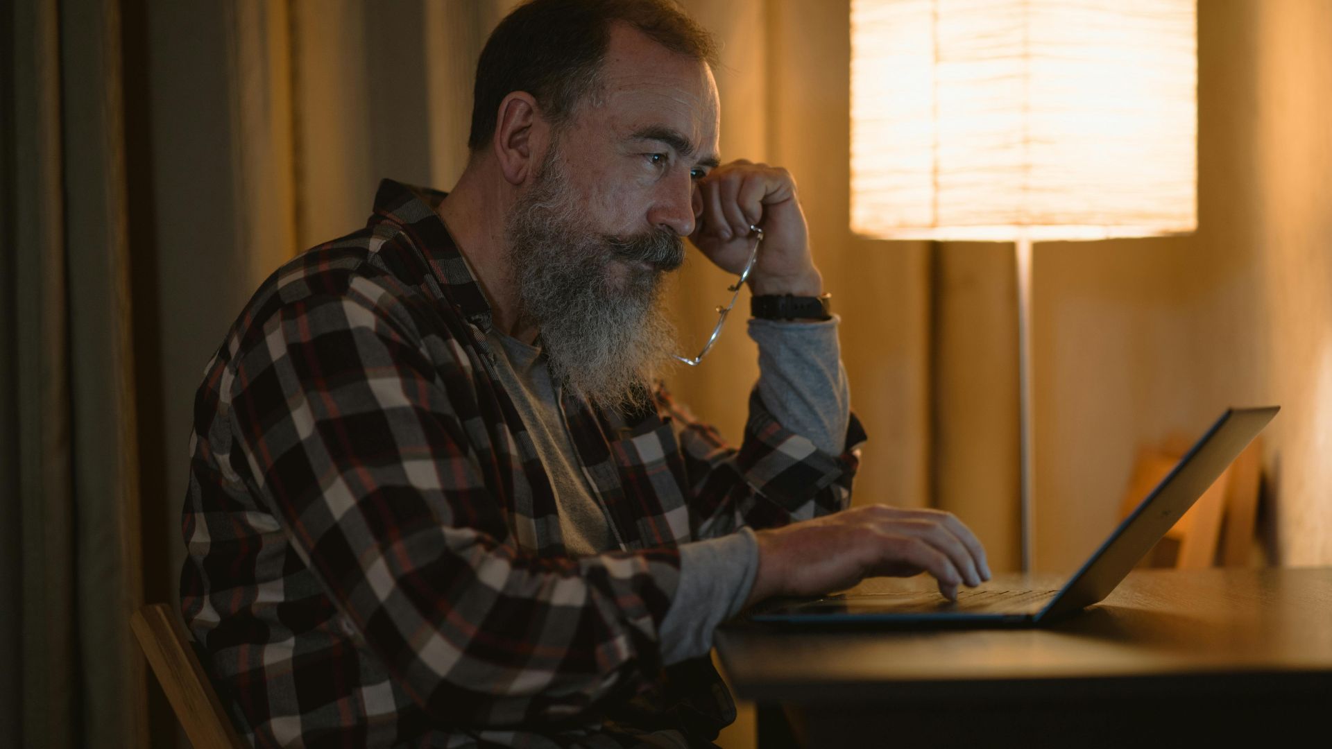Elderly man with a beard working late on laptop under warm lamp light indoors.