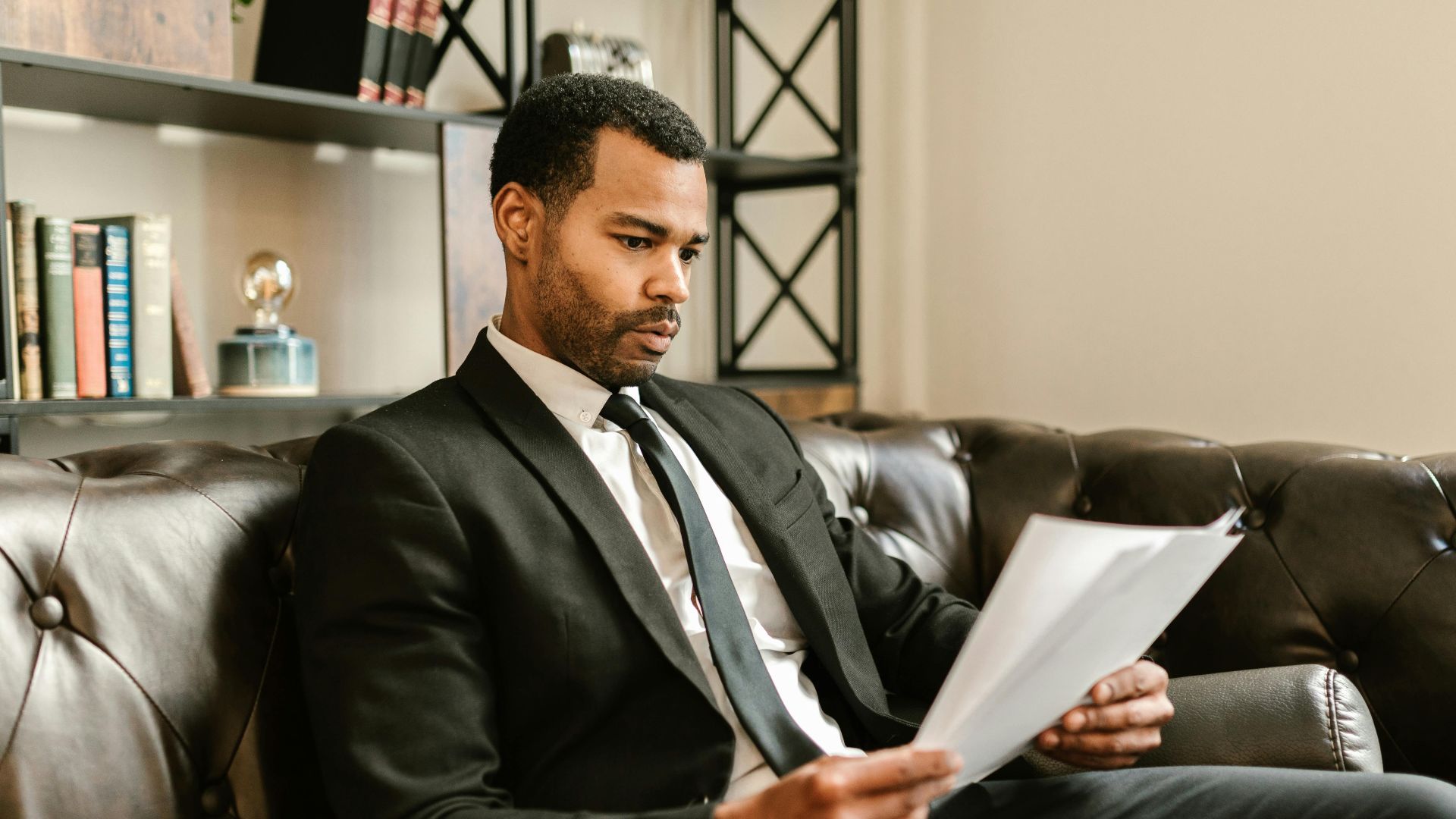 Businessman in suit reading papers on a leather sofa in a modern office setting.