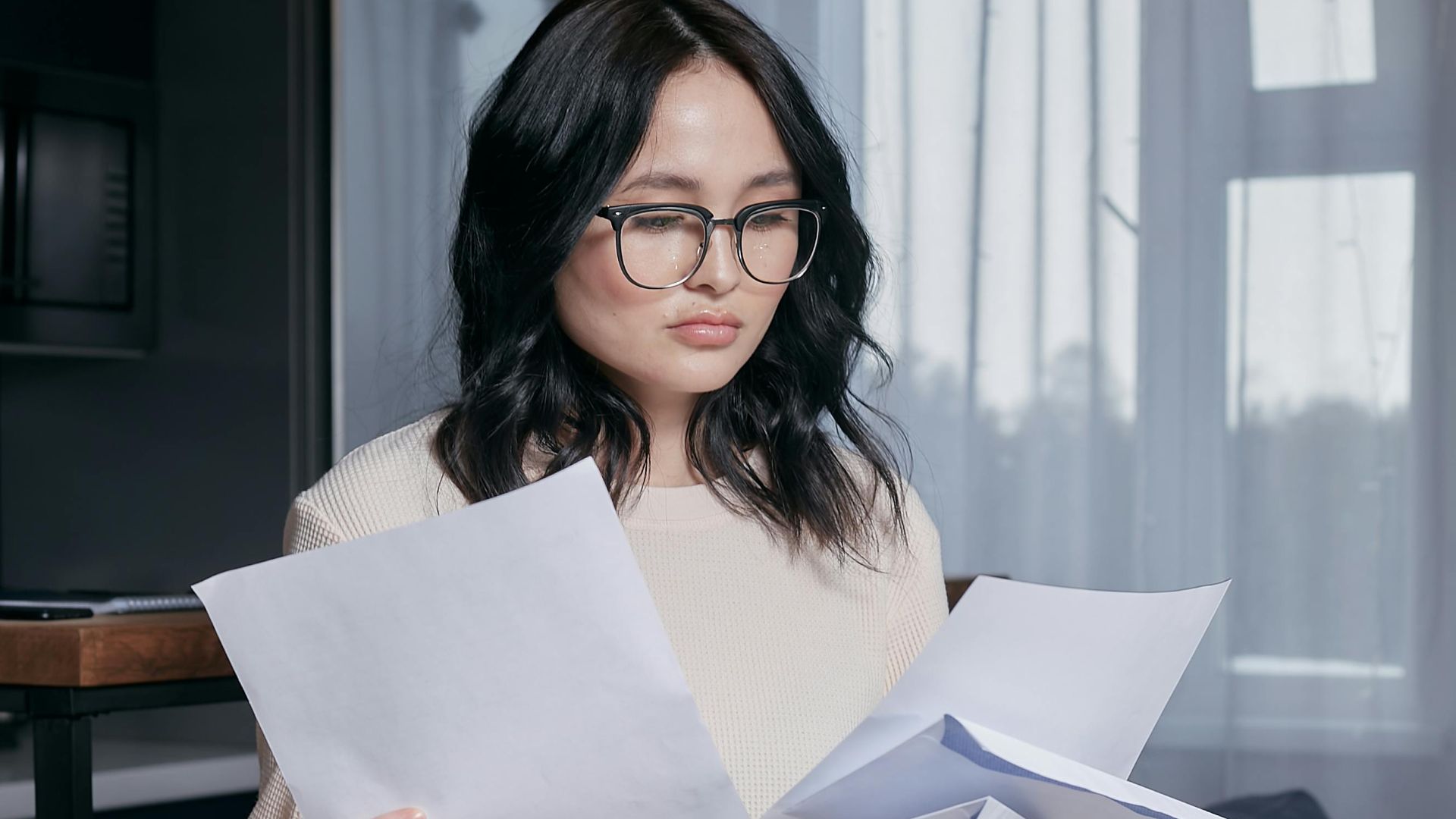 A woman sits indoors, reviewing documents with focused attention near a window.