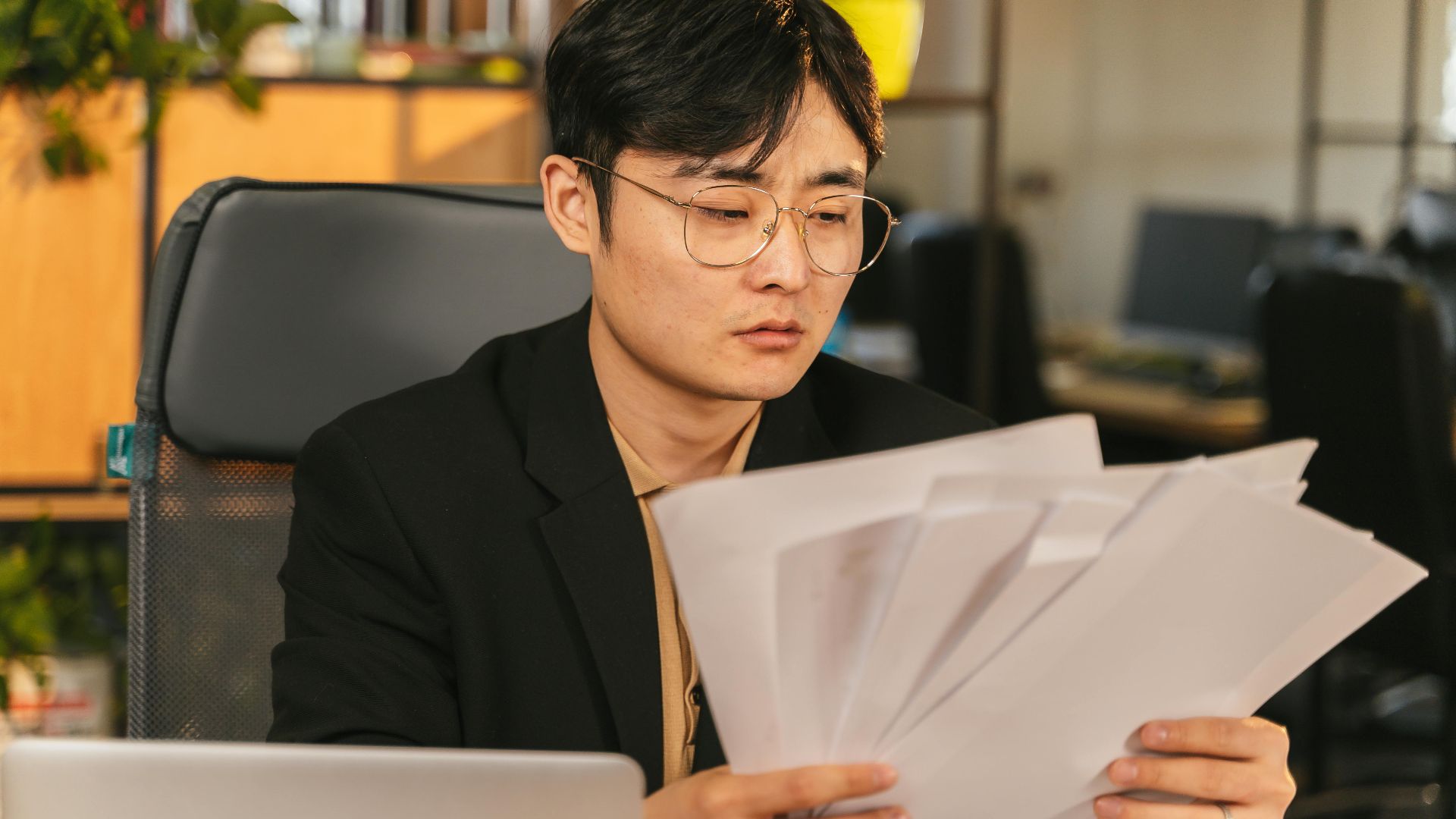 Asian man in glasses and suit analyzing papers at his desk in a modern office.