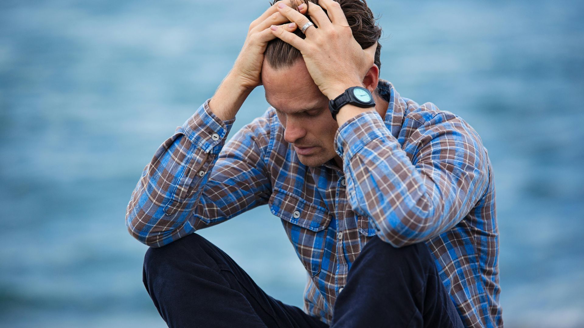 A man in a plaid shirt sits by the water looking distressed, symbolizing stress.