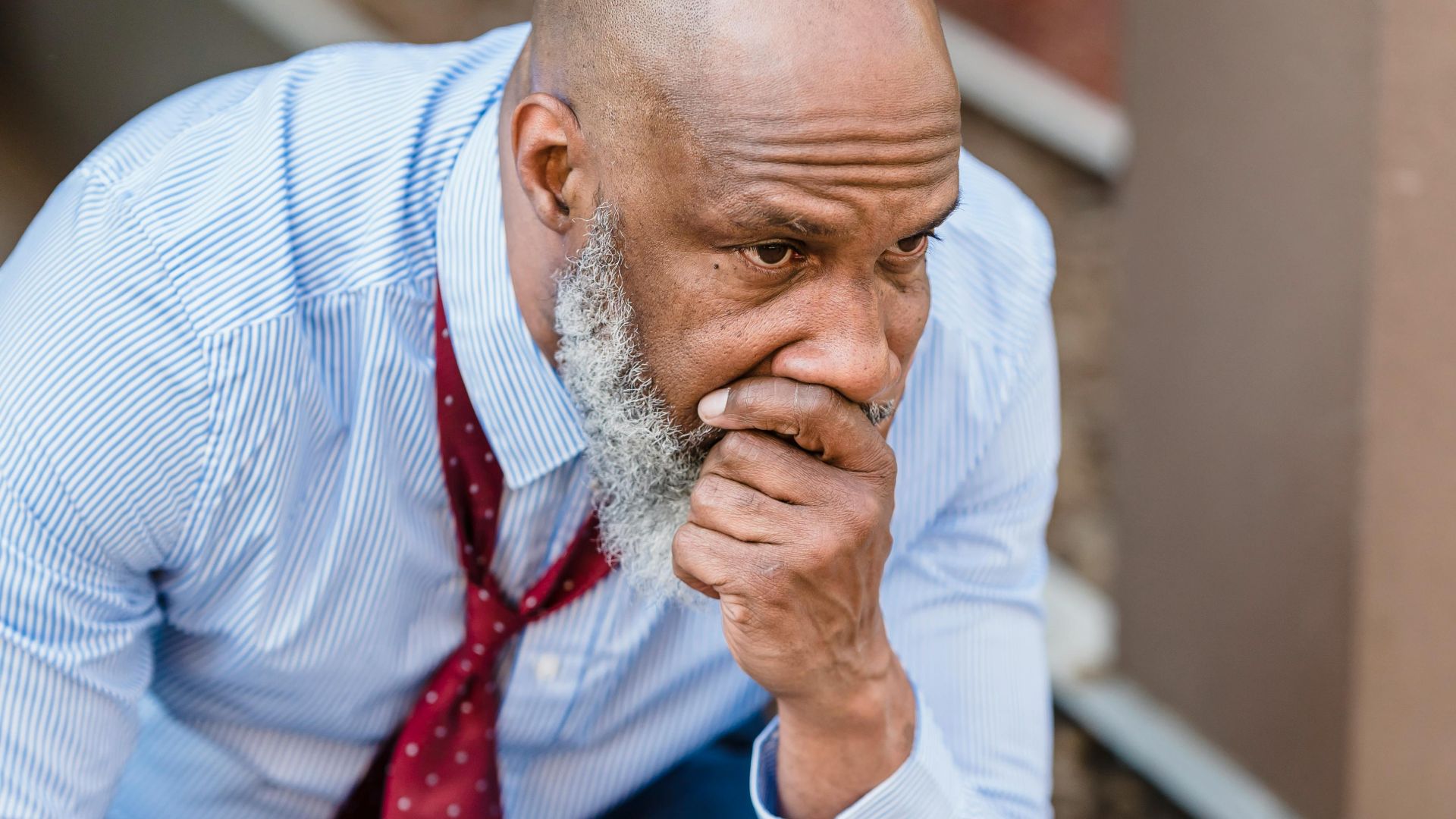 Pensive unhappy African American male worker in formal clothes covering mouth while sitting on street of city after hard workday