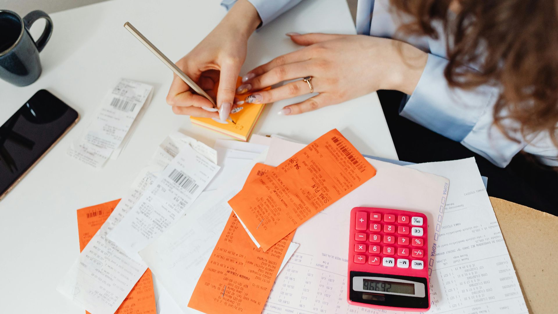 A woman writes down financial details using a calculator, receipts, and documents.