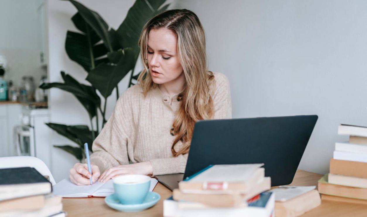 Pensive businesswoman writing information in notebook near laptop and coffee