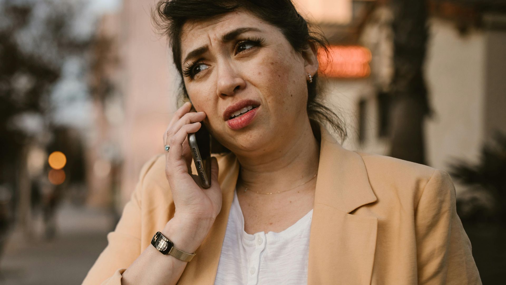 Woman in beige jacket looks concerned while making phone call outdoors.
