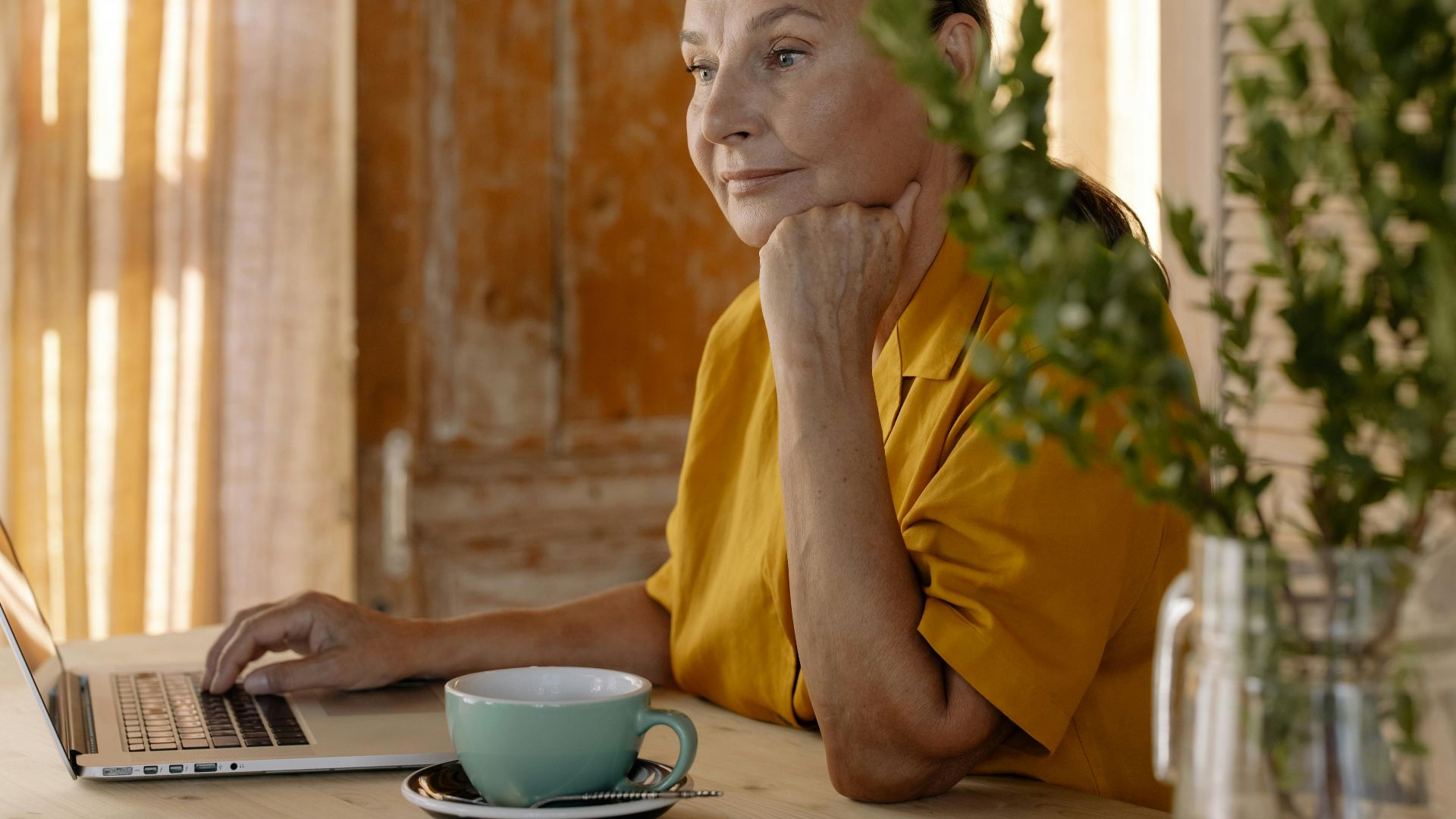 Elderly woman sitting at a table using a laptop, relaxing in a cozy home setting.