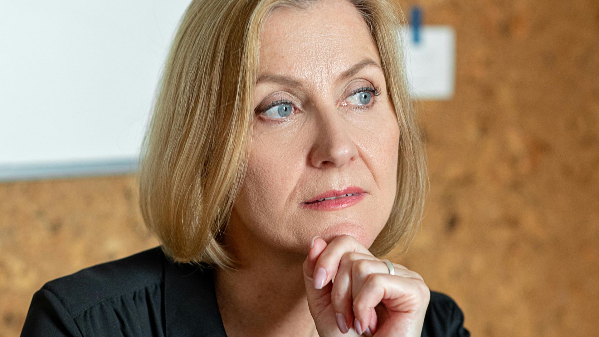 Mature businesswoman in black attire thinking at a wooden desk in a modern office.
