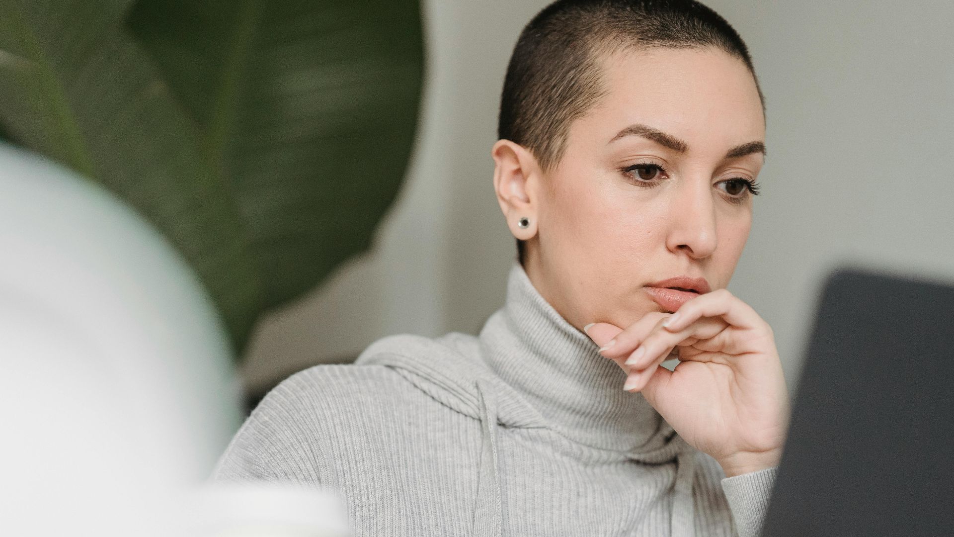 Thoughtful female with short hairstyle looking at netbook screen and touching chin in light room