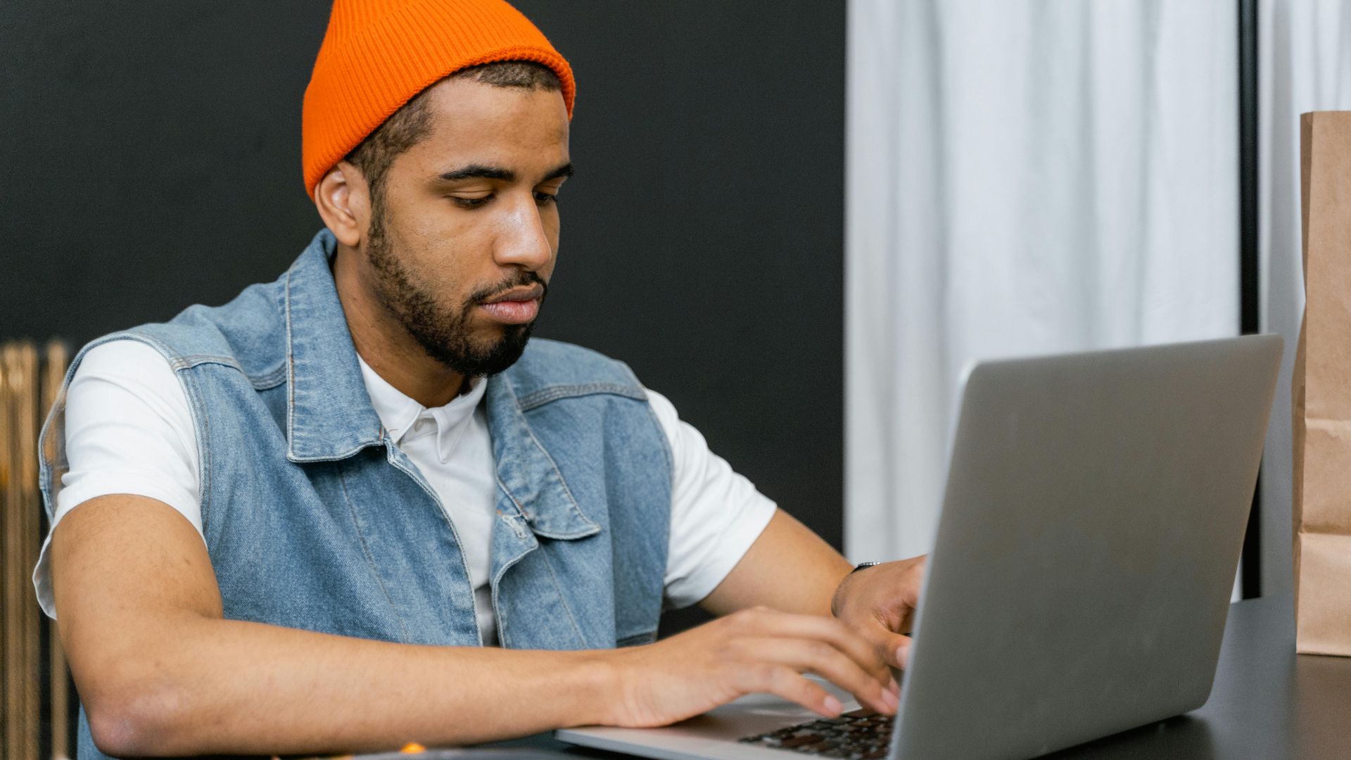 Young man in orange beanie working intently on a laptop at a desk.