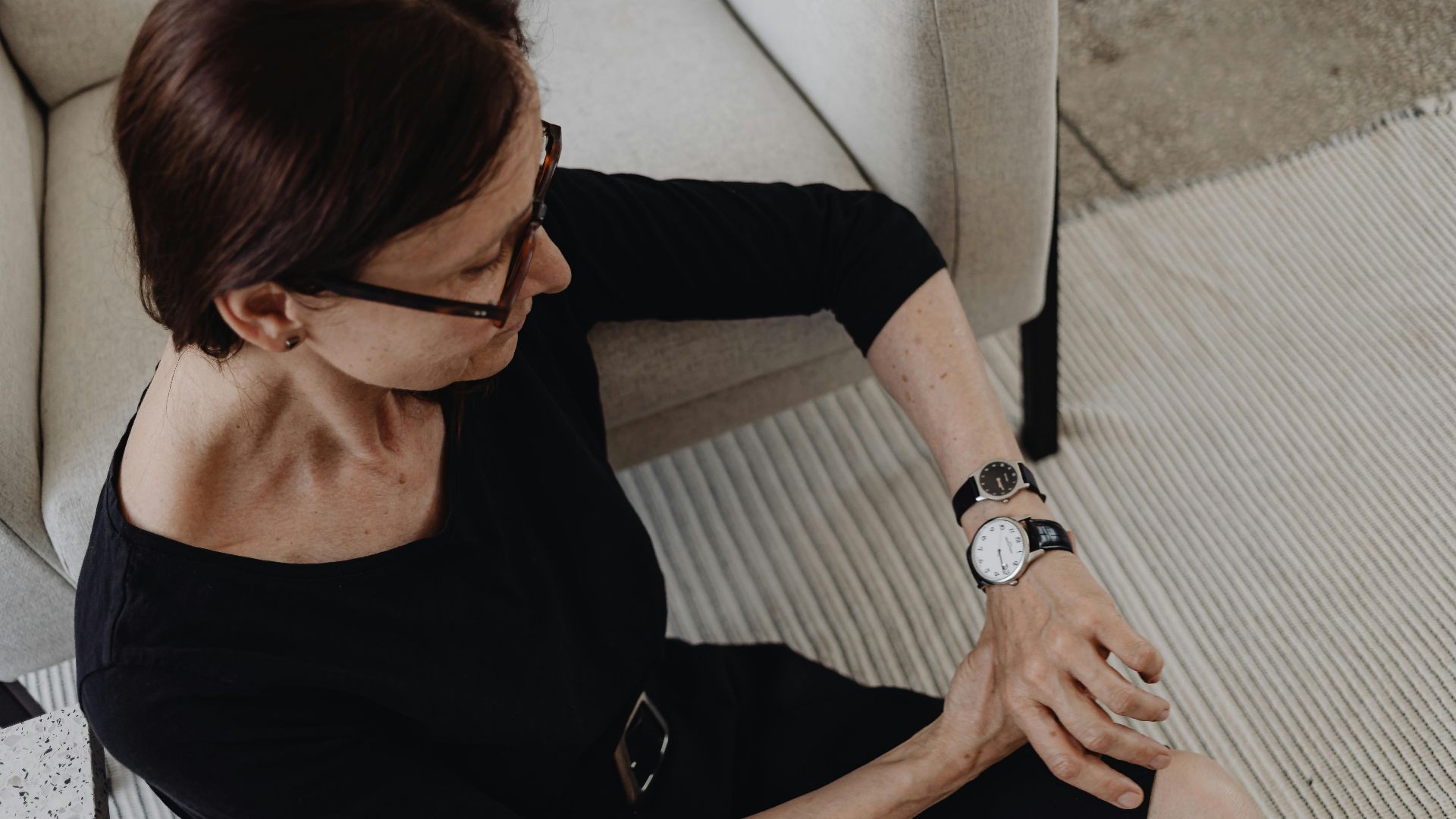 Mature woman in black dress sitting indoors, checking her wristwatch, expressing contemplation.