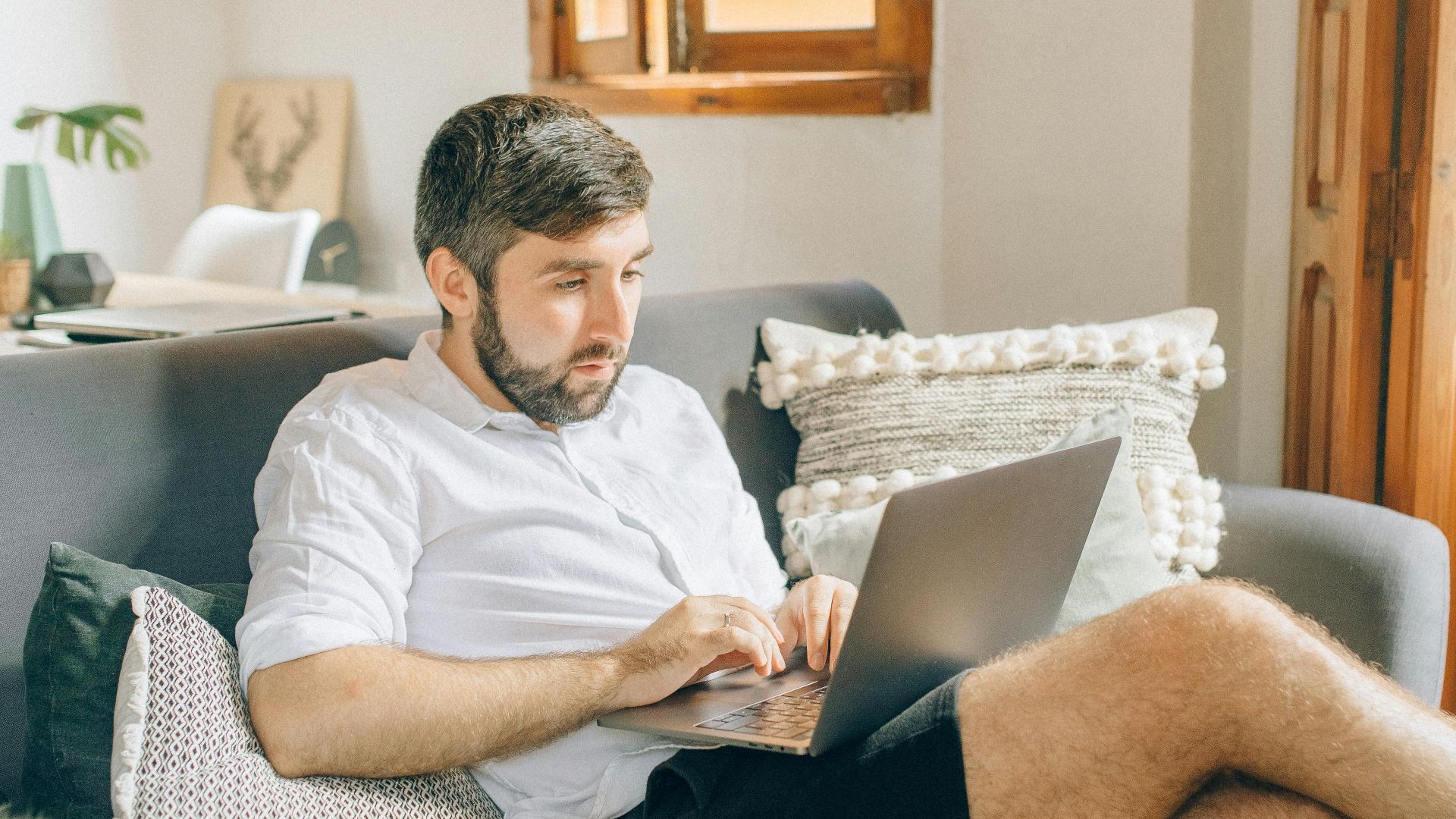 Adult man sitting on couch using laptop, working remotely from cozy home setting.