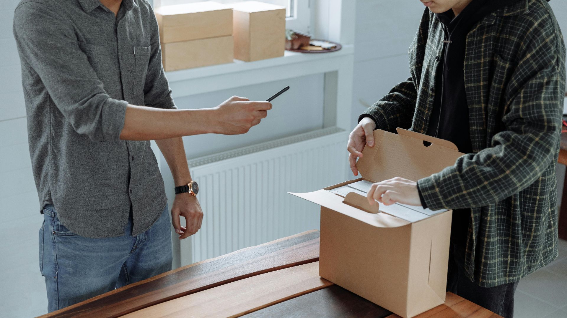 Two adults unpack and photograph a cardboard box using a smartphone indoors.