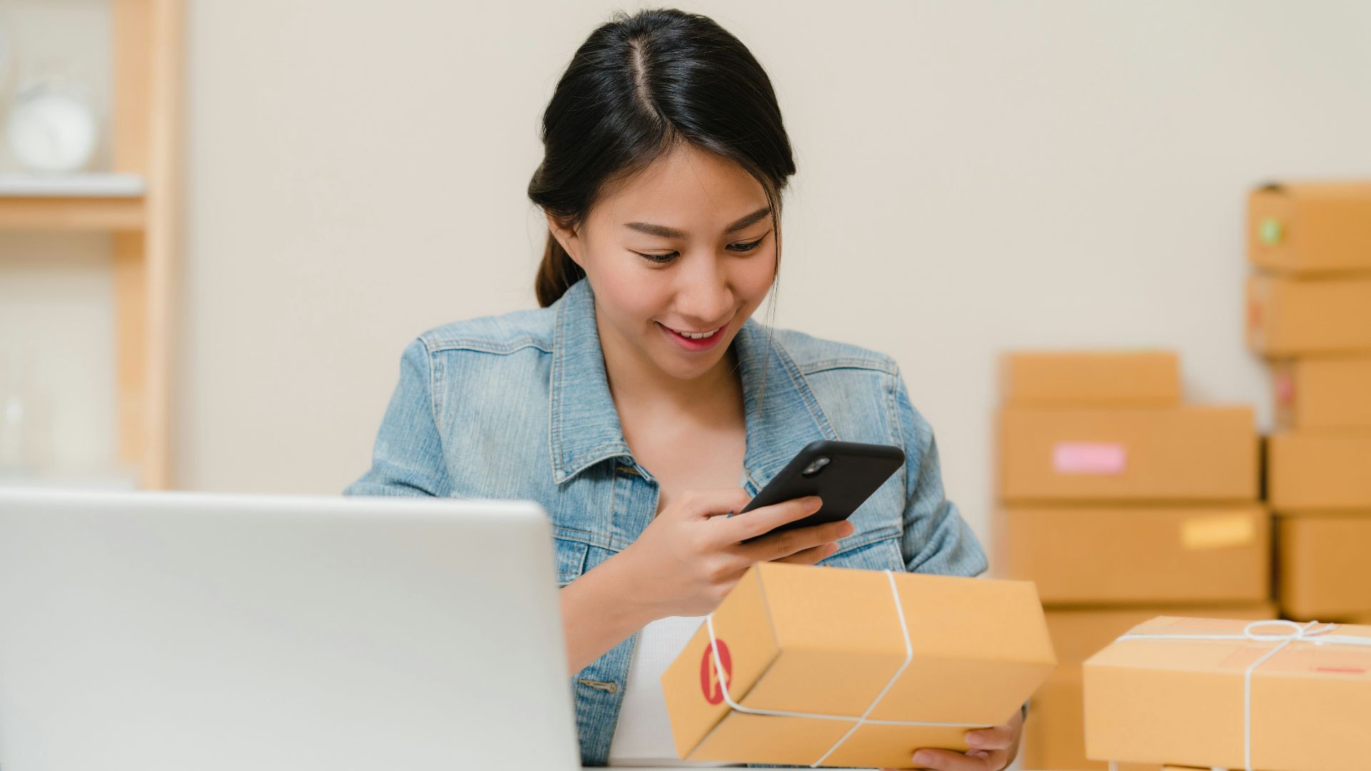 Woman checking package with phone near laptop and boxes