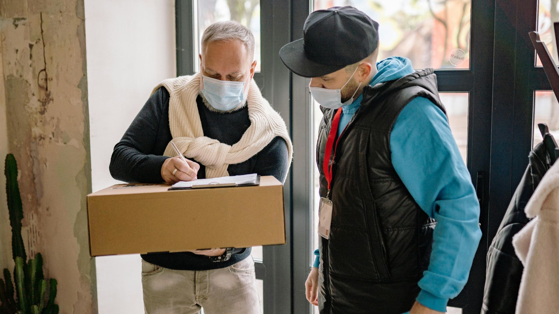 Elderly man signs for a parcel while a delivery man wearing a mask observes indoors.