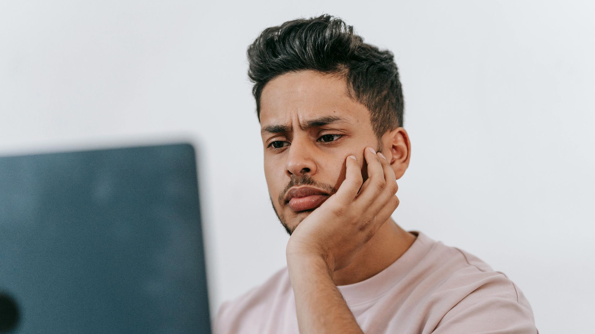 Serious young ethnic male in t shirt browsing netbook and leaning on hand in light room