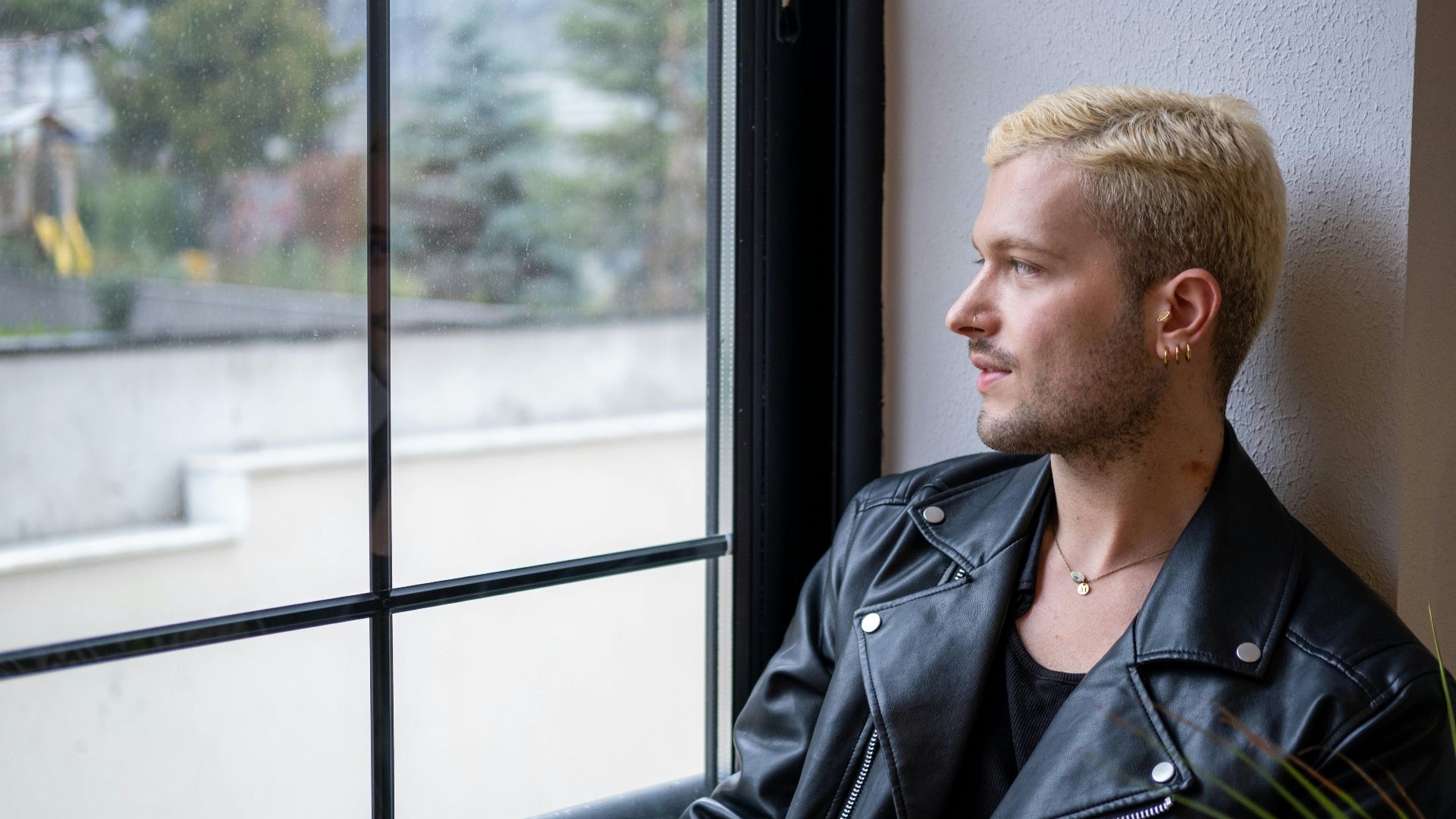 A man with blonde hair and a leather jacket gazes thoughtfully out the window indoors.