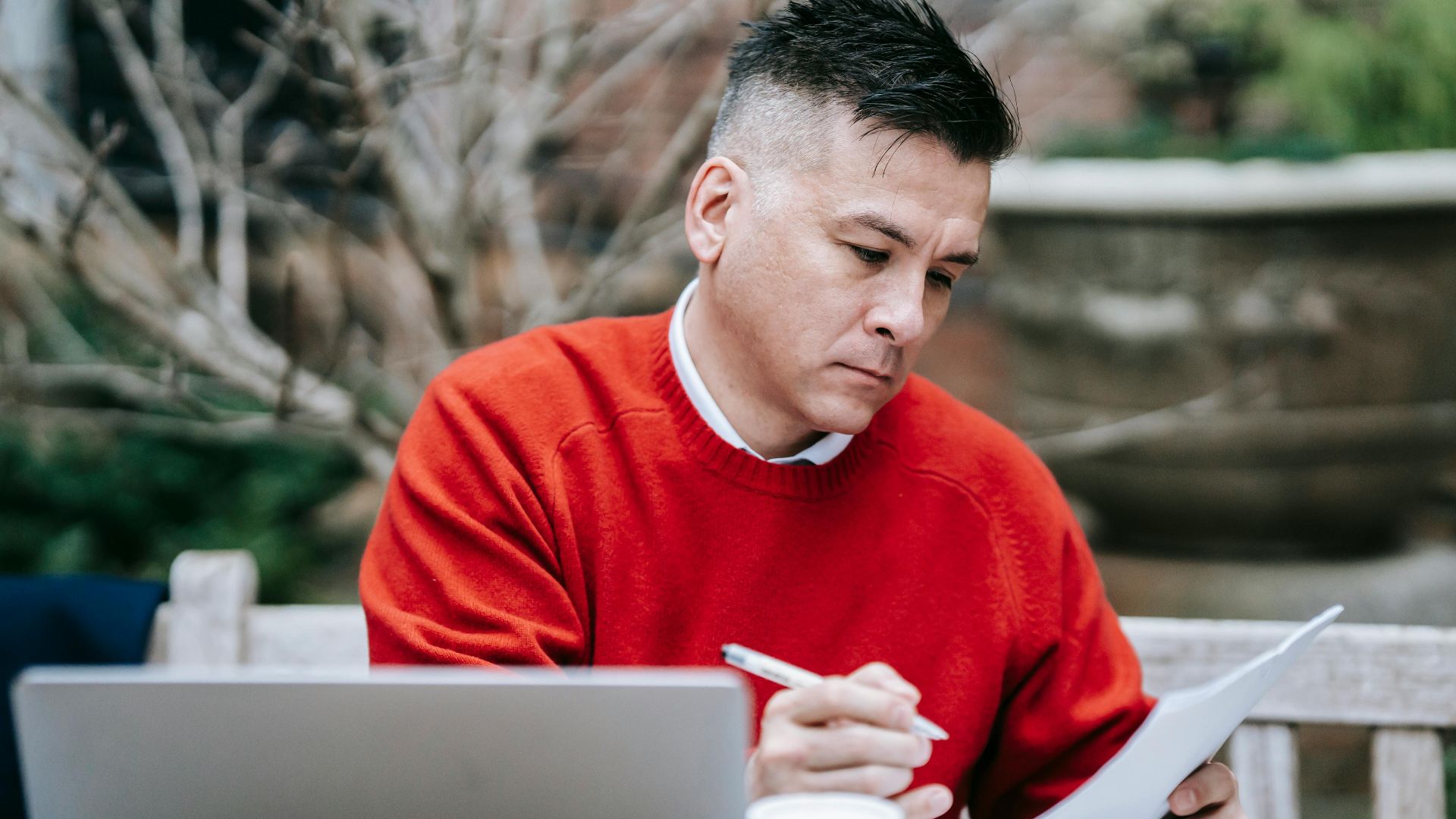 A man in a red sweater concentrating on paperwork while working remotely with a laptop outdoors.