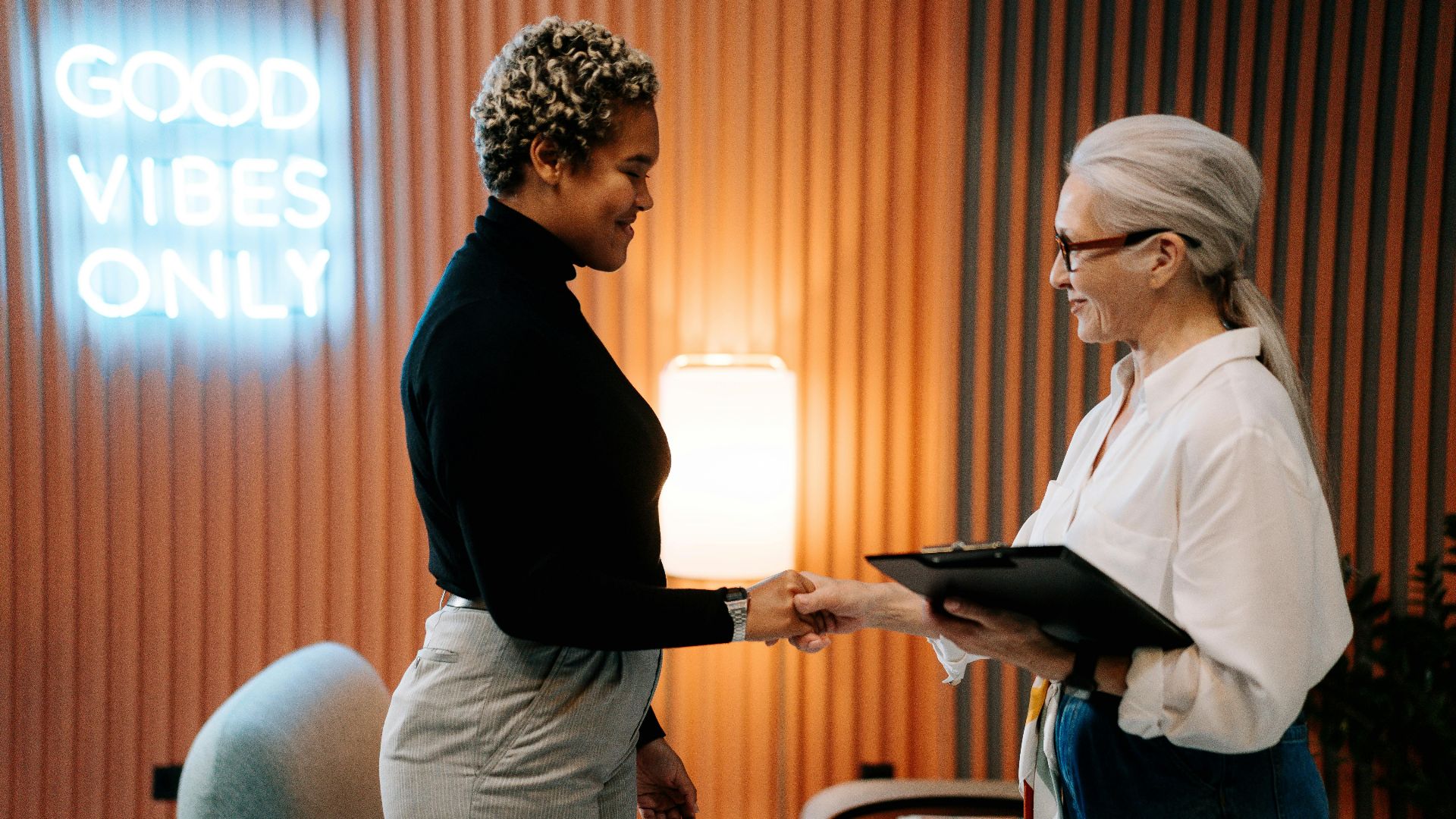 Two diverse businesswomen in a stylish office shaking hands under a 'Good Vibes Only' sign.