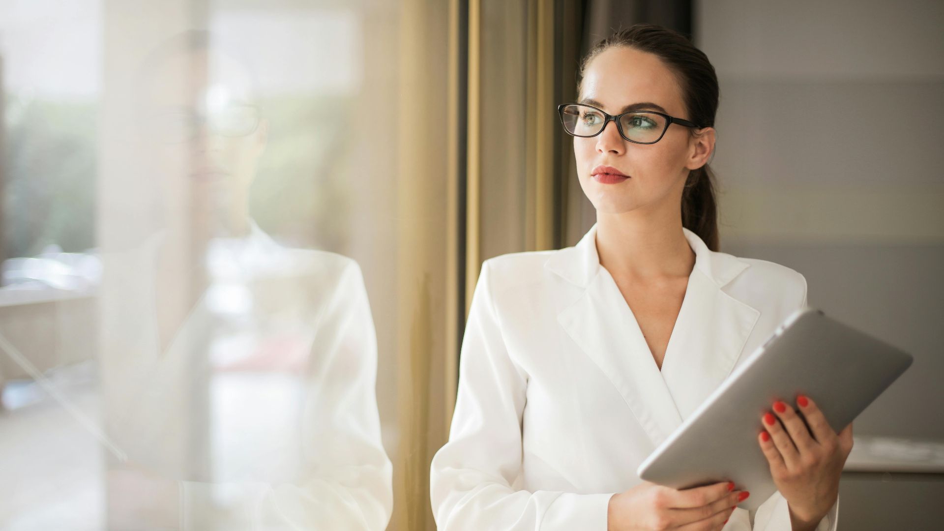 Businesswoman in formal attire thinking beside a window with a tablet.