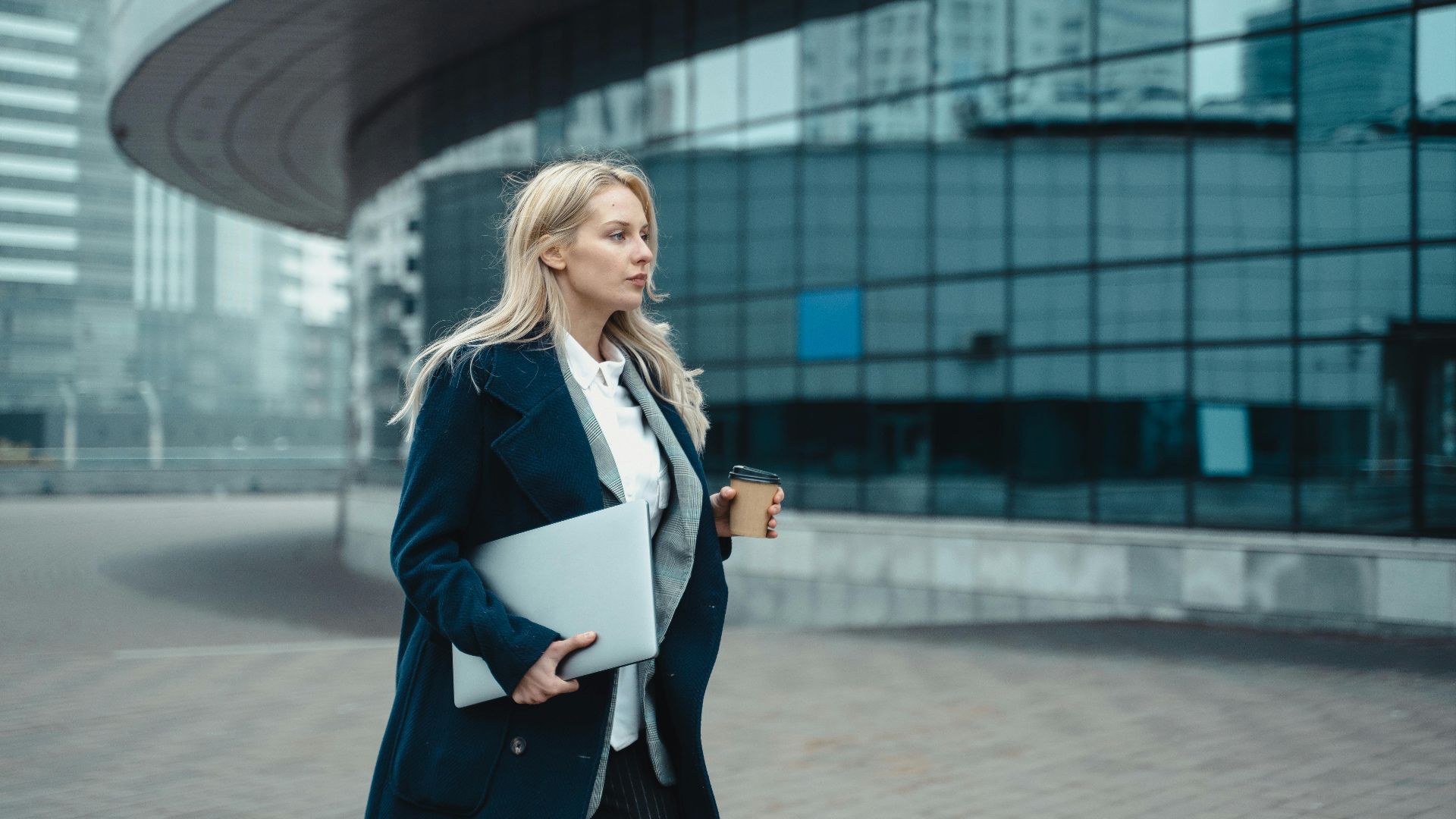 Confident businesswoman walking outside modern office building holding laptop and coffee.