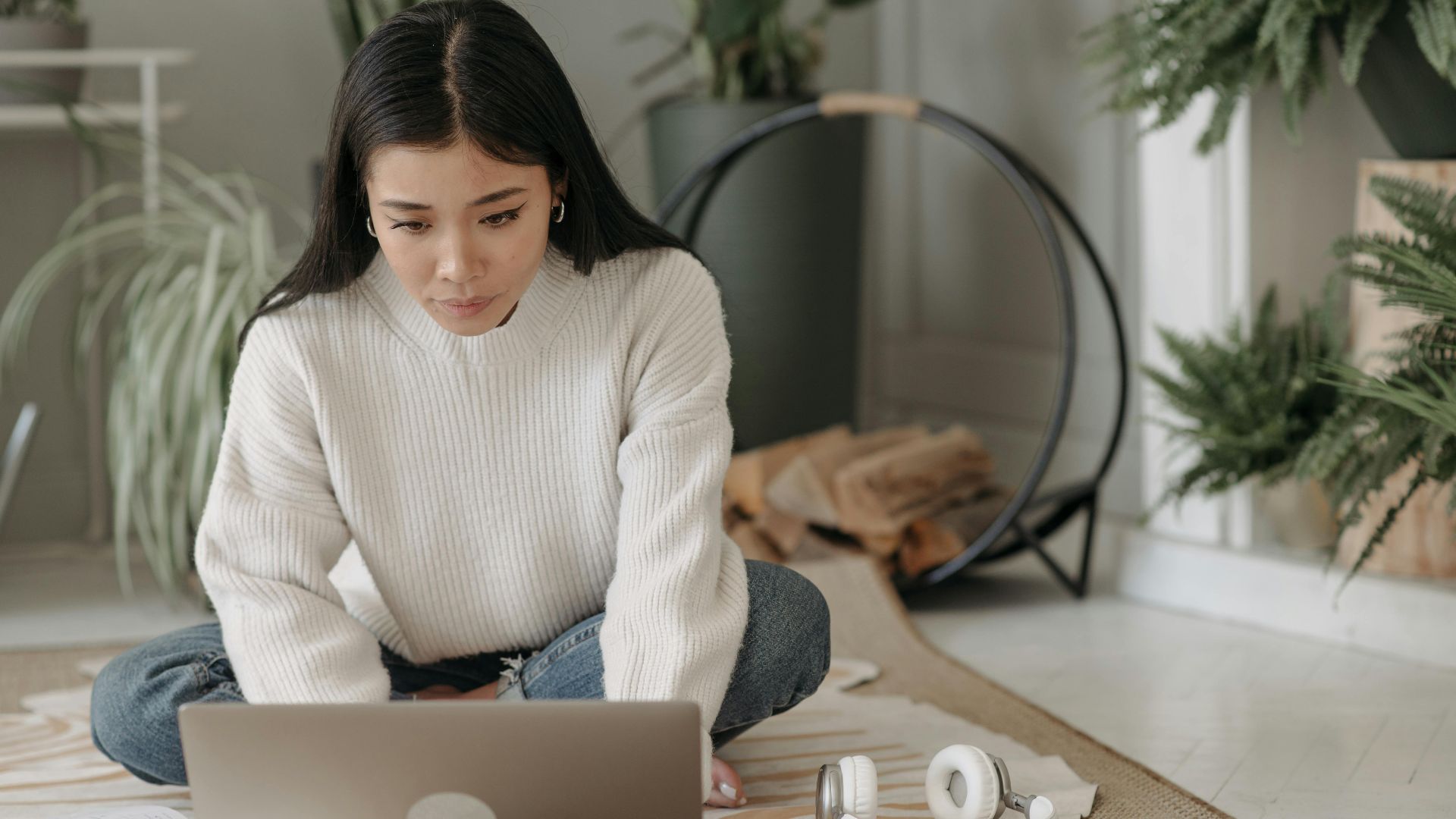 Woman sitting on carpet using laptop at home, surrounded by plants, focused on remote work.