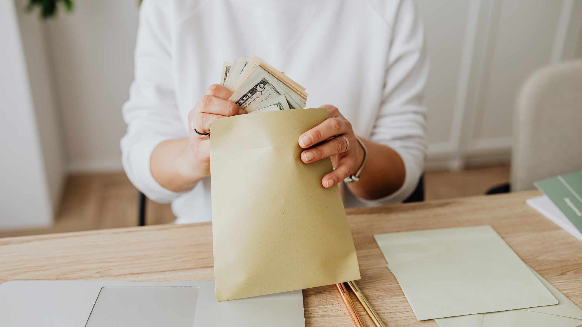 Close-up of person holding envelopes with cash at a wooden desk indoors.