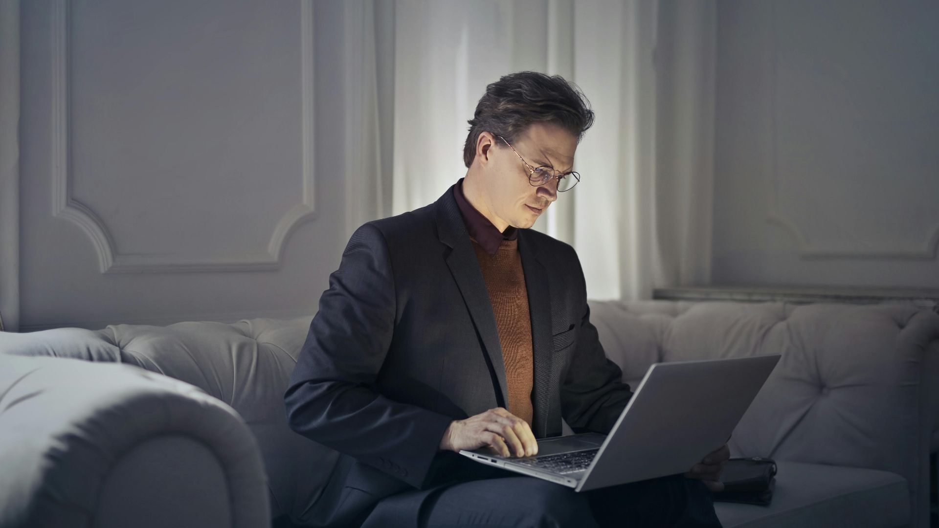 Professional man sitting on sofa using laptop for remote work in a stylish indoor setting.