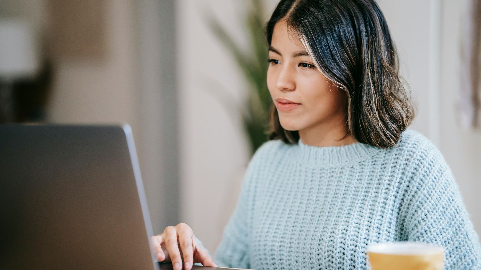 Confident young Latin American woman in casual clothes working remotely on netbook at table near mug with coffee in light apartment with plant with green leaves near wall