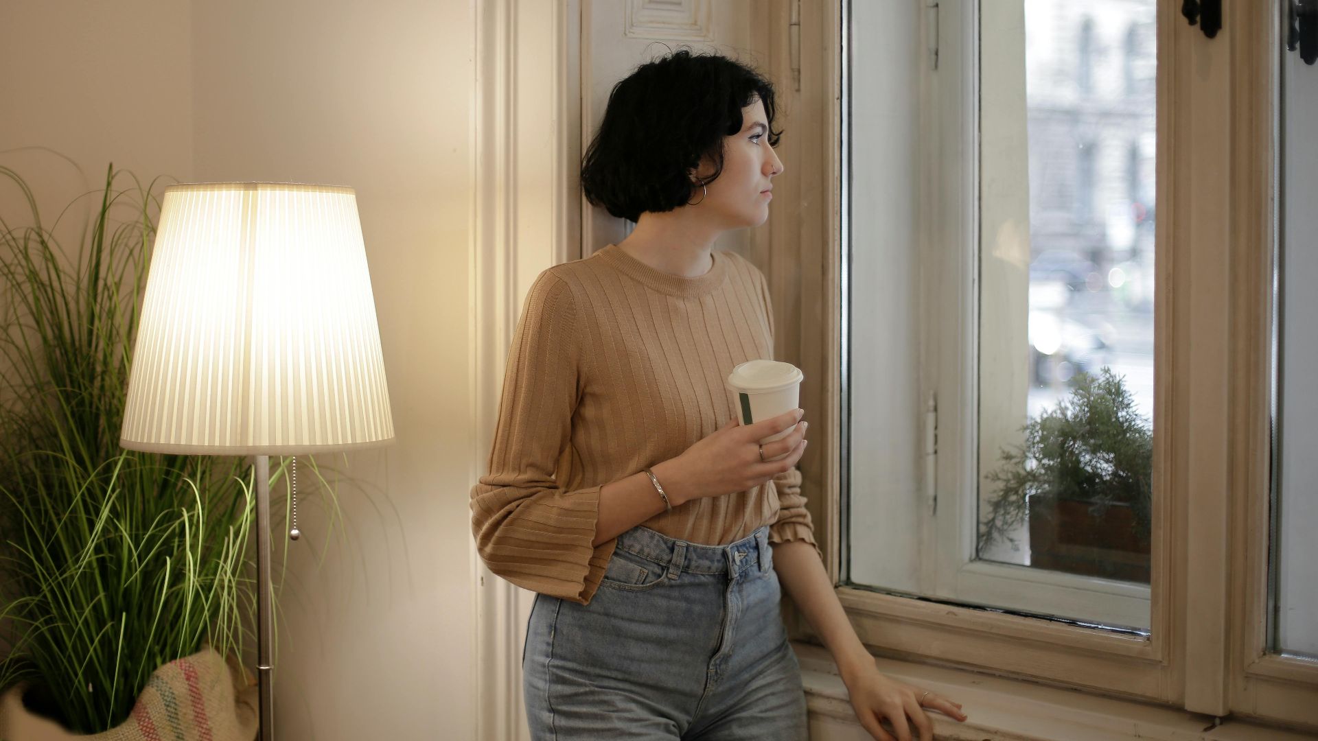 Young woman holding a coffee cup, gazes thoughtfully out the window in cozy room.