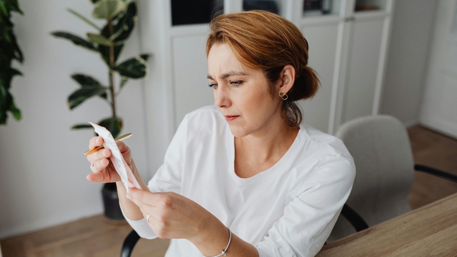 A concerned adult woman examines a receipt while sitting at her office desk, indicating financial scrutiny.
