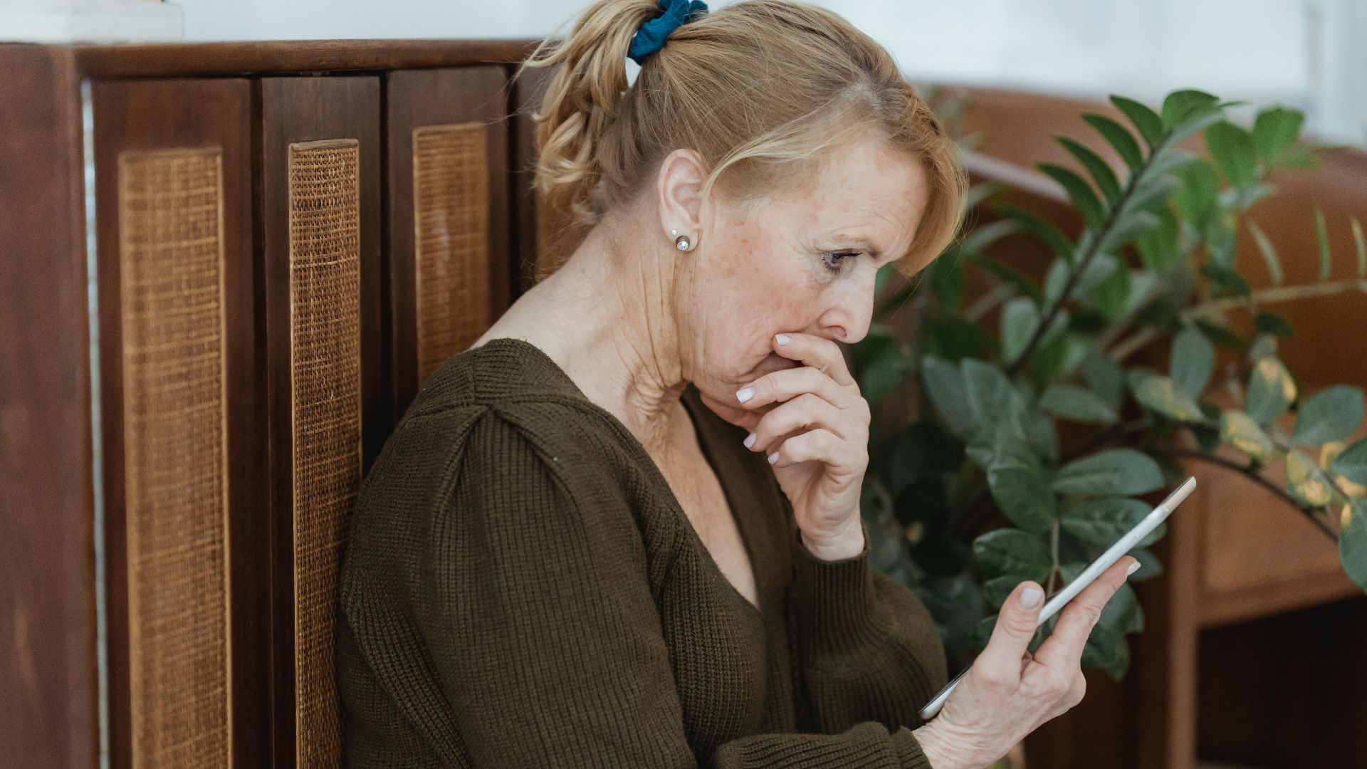 Side view of surprised elderly female with cellphone covering mouth against cabinet and plant at home