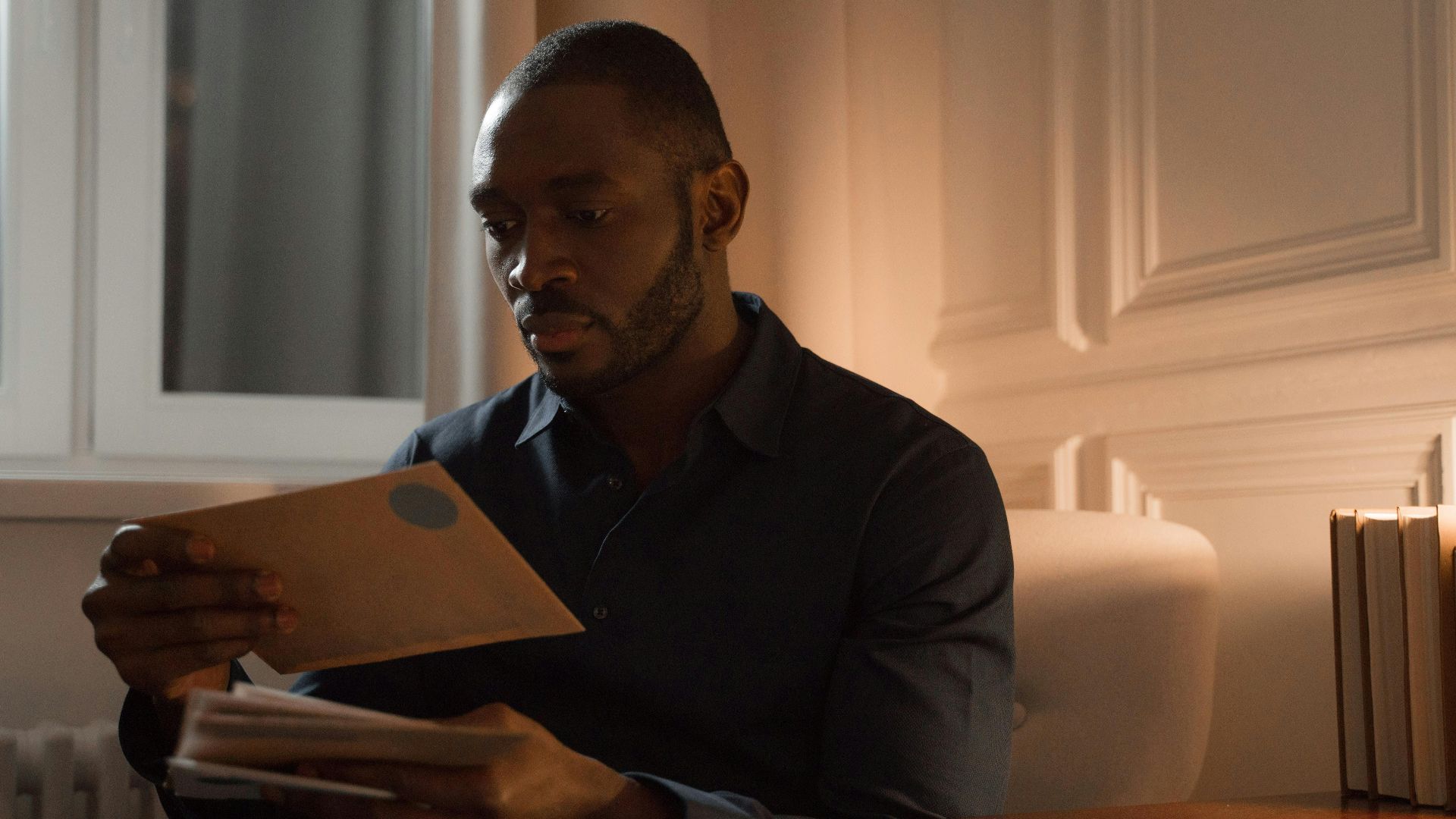 A man with a mustache reads letters while sitting in a warmly lit home interior.