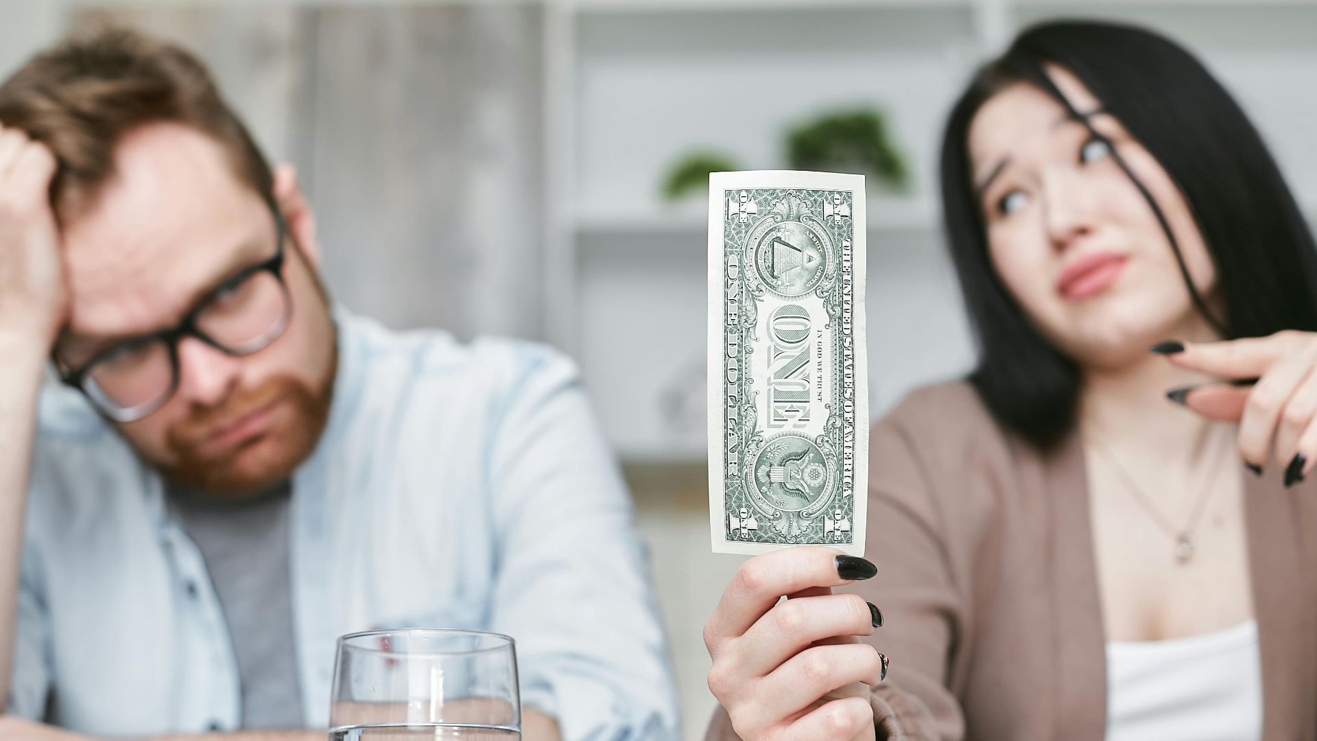 An adult interracial couple expressing concern about finances as they hold a one dollar bill indoors.