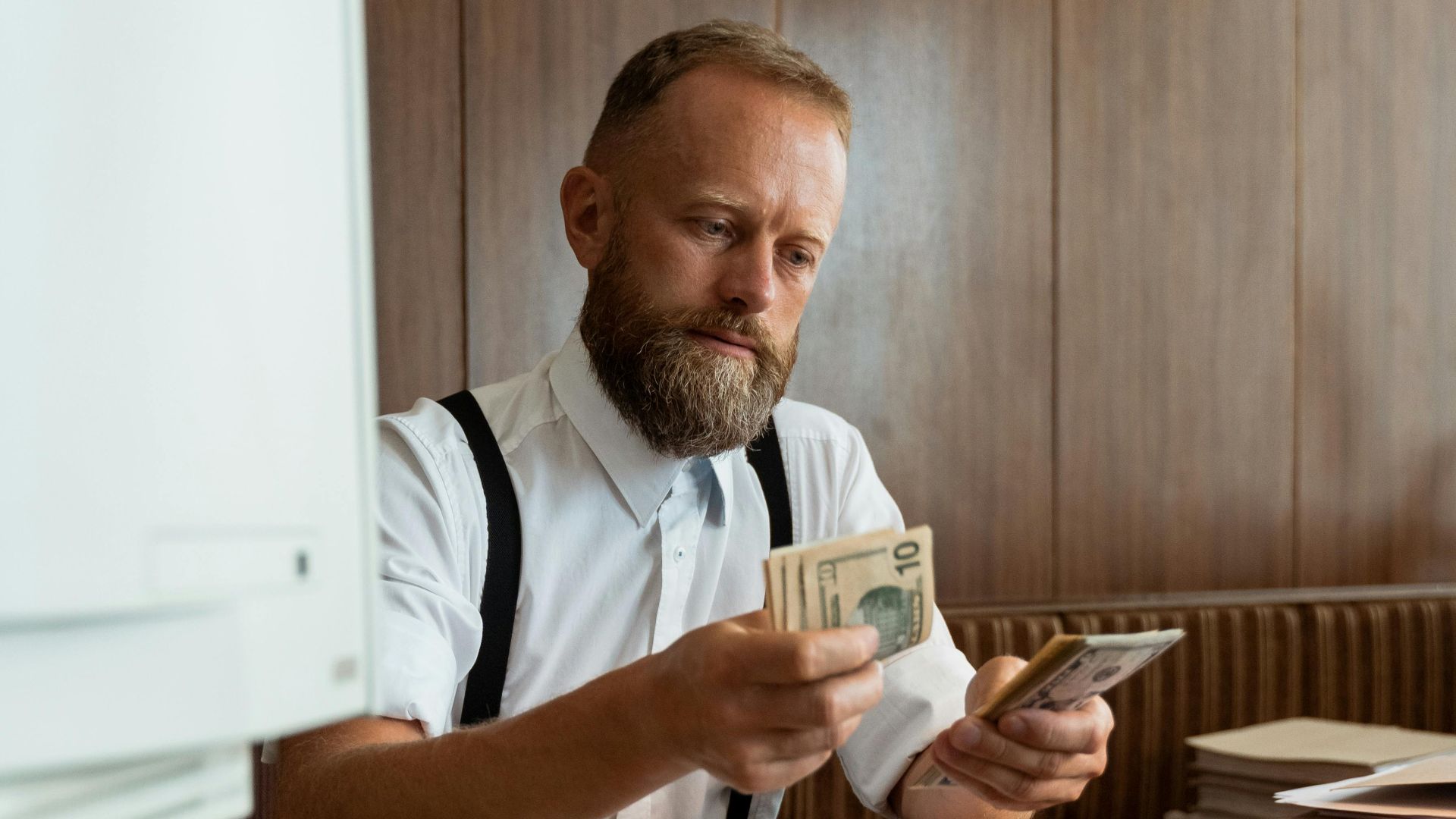 A bearded man counting cash in a vintage office with an old computer.