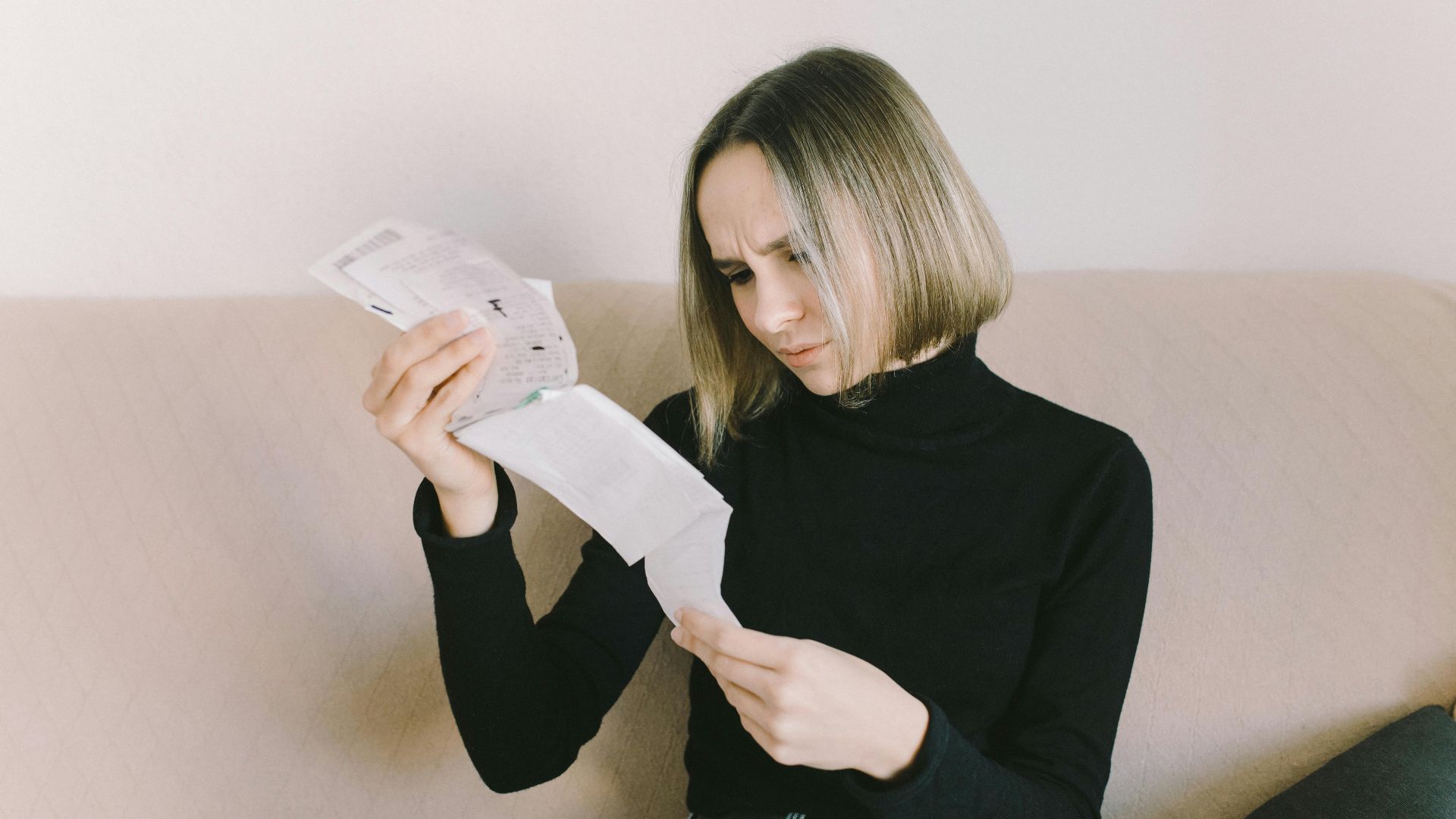Concerned woman in black sweater examining bills on beige sofa indoors.
