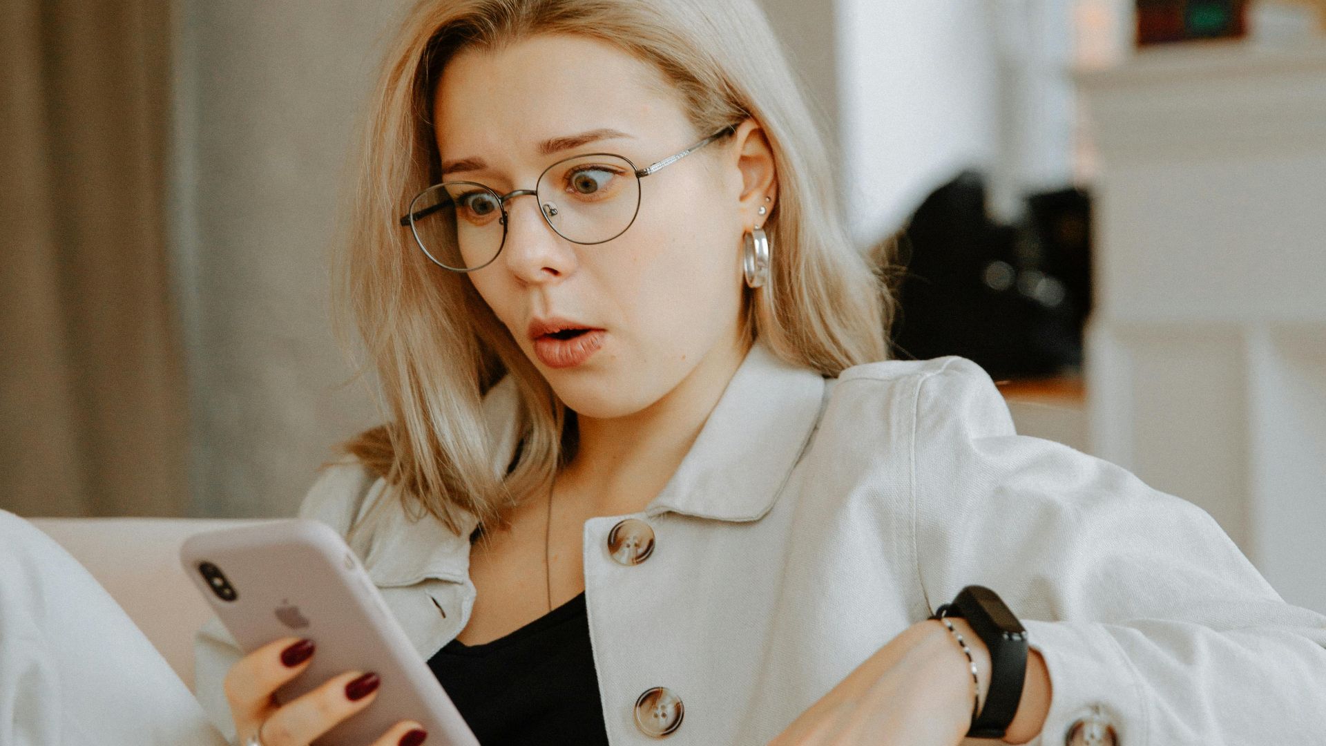 Surprised woman with glasses reacts to news on her phone while relaxing indoors.