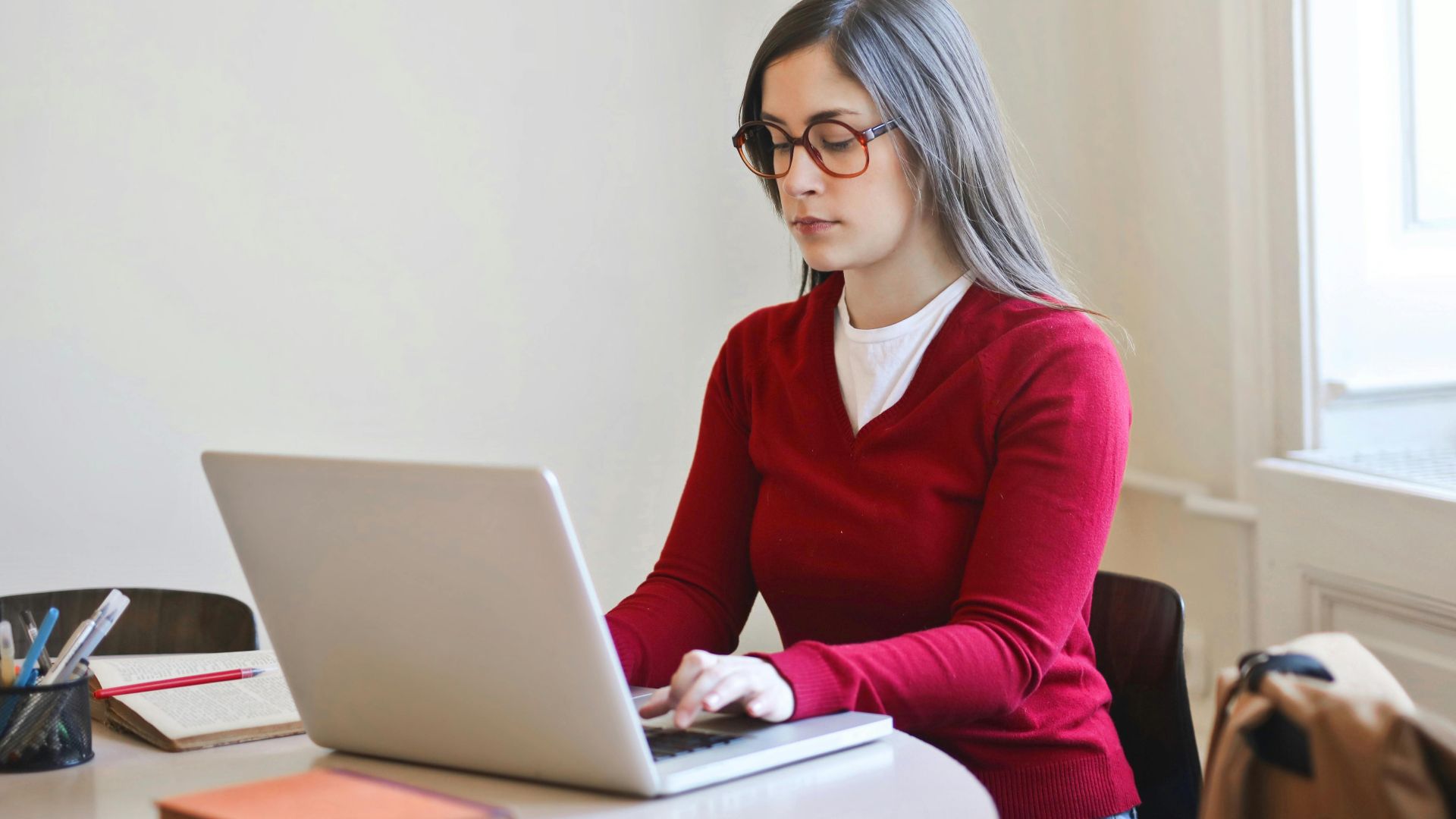 Young woman in red sweater working remotely on a laptop at a table indoors.