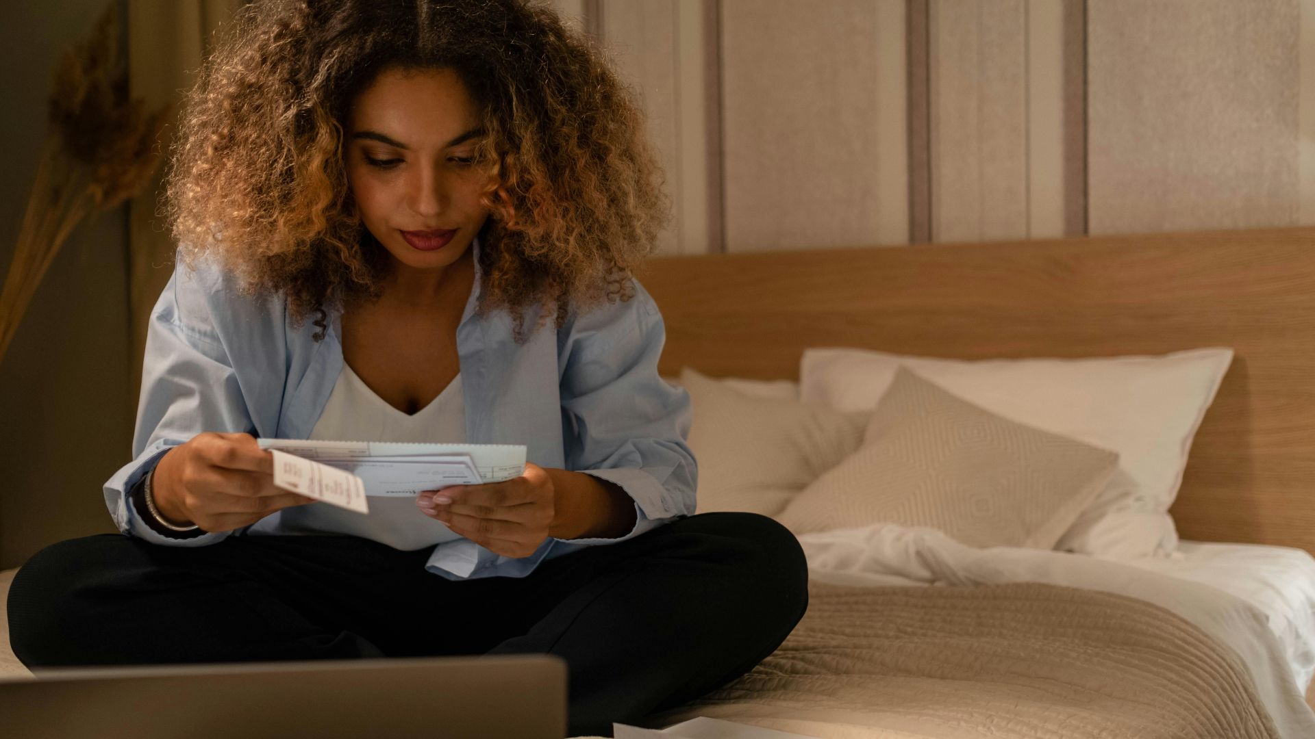 Woman sitting on bed reading documents with laptop, highlighting home office setting.