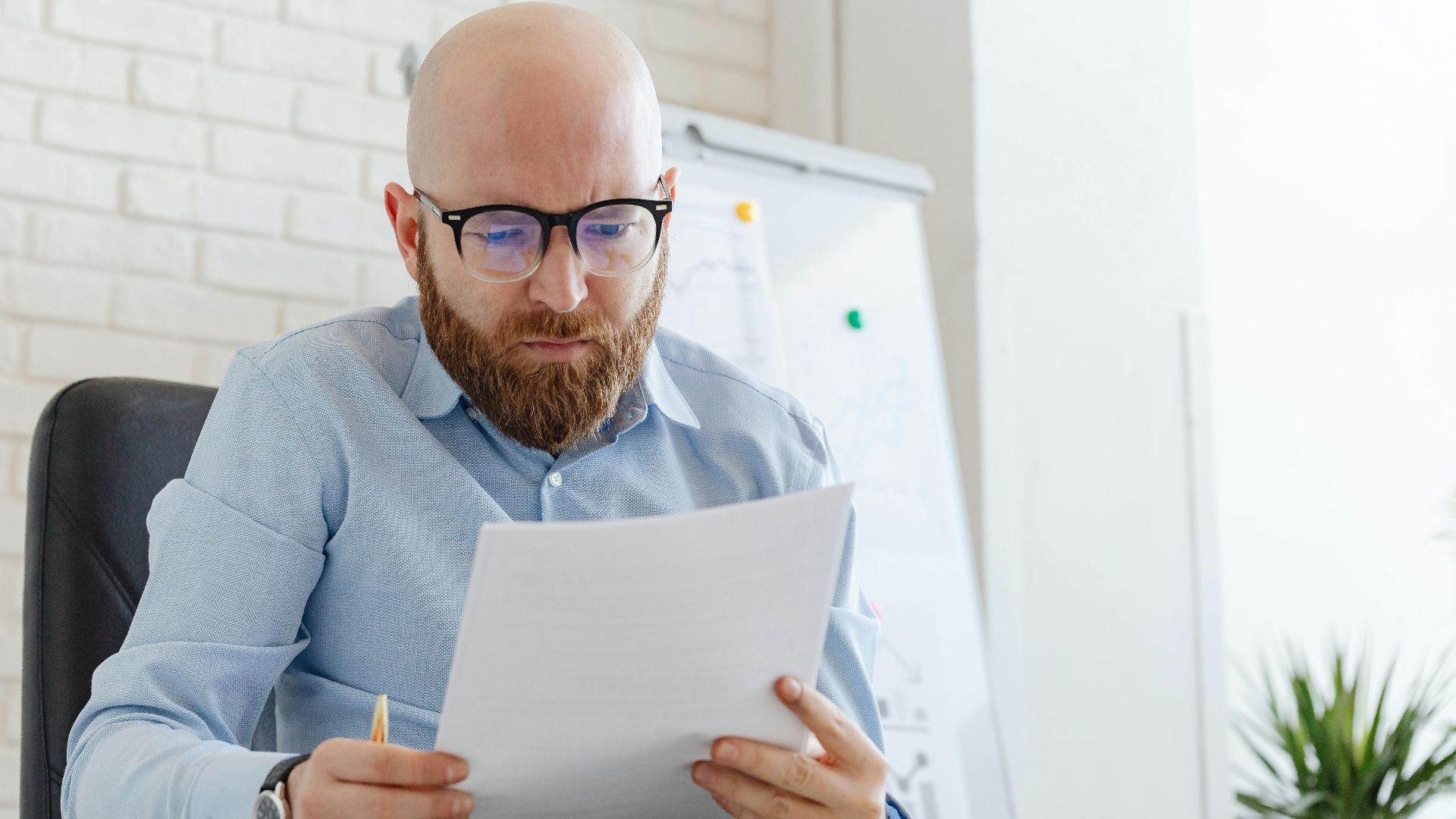 A businessman with a beard and eyeglasses reviewing documents in an office setting.
