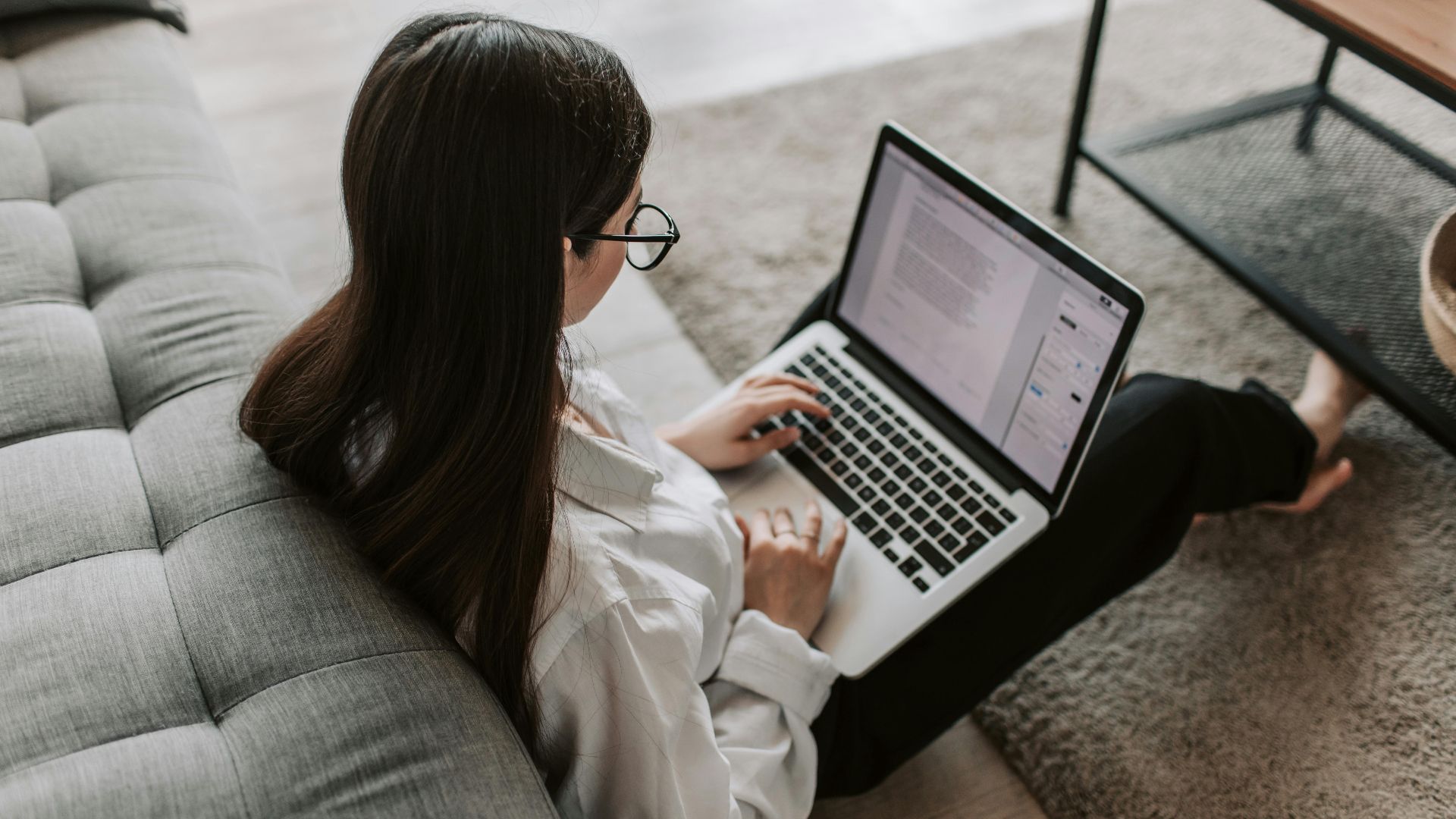 Woman working remotely with a laptop on the floor next to a sofa, enjoying comfortable home office setup.