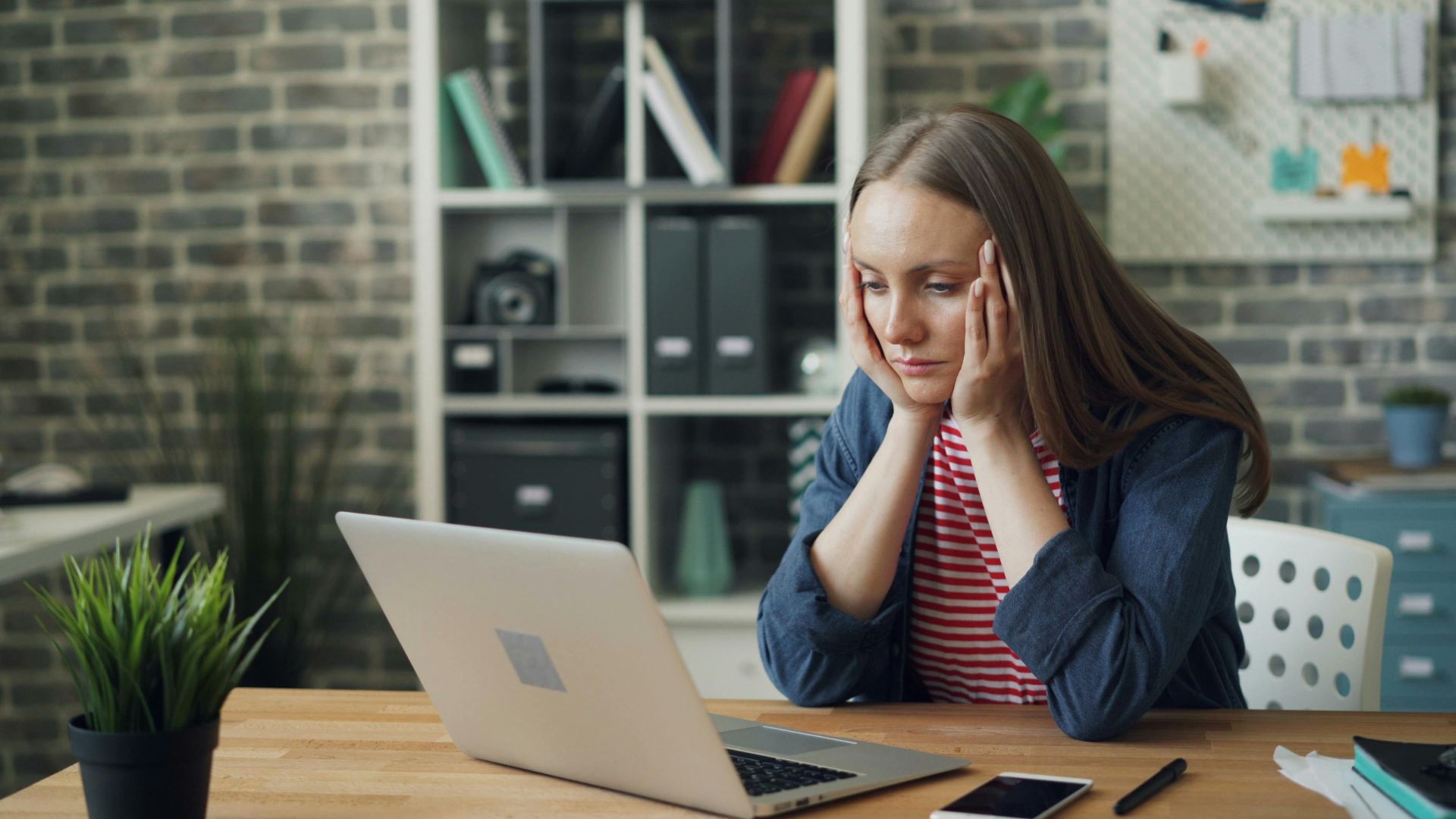 A woman appears stressed while working on a laptop in her modern office.