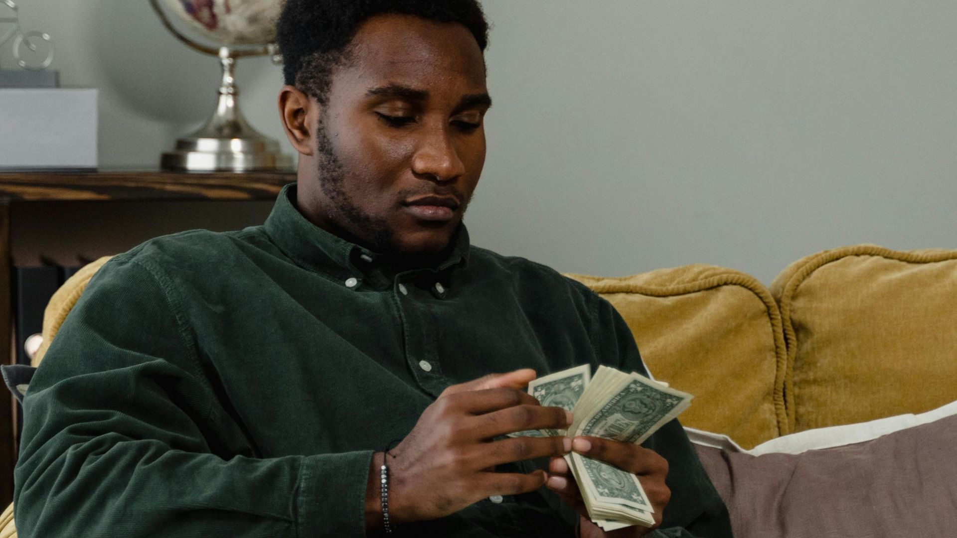 A man sitting on a couch counting cash notes in a cozy indoor setting with a globe in the background.