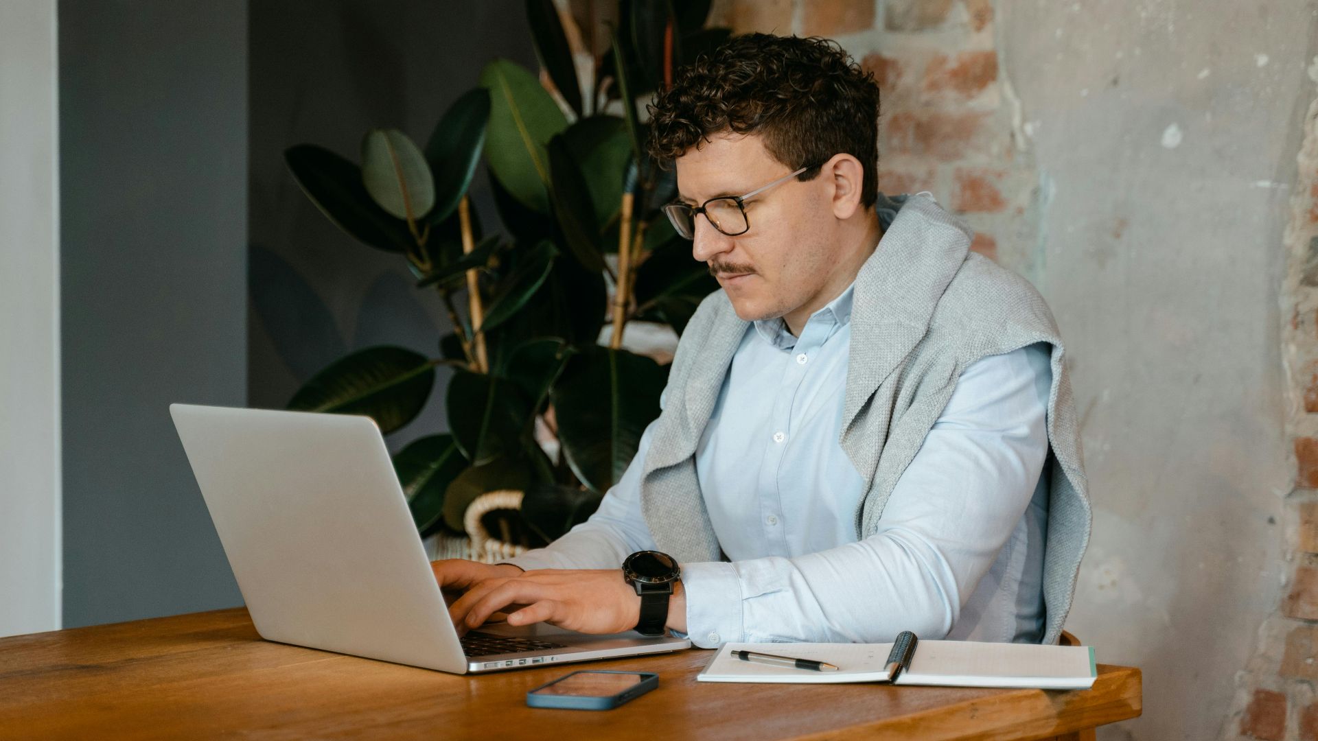 Adult man using a laptop at a wooden table in a cozy workspace with exposed brick walls.