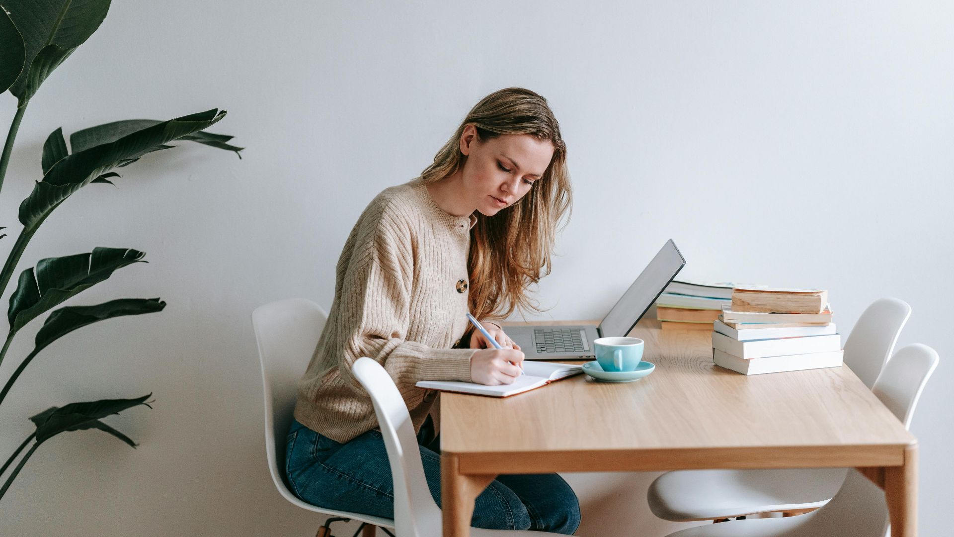 Side view of concentrated female freelancer taking notes in notepad while working at table with netbook in light room with green plant