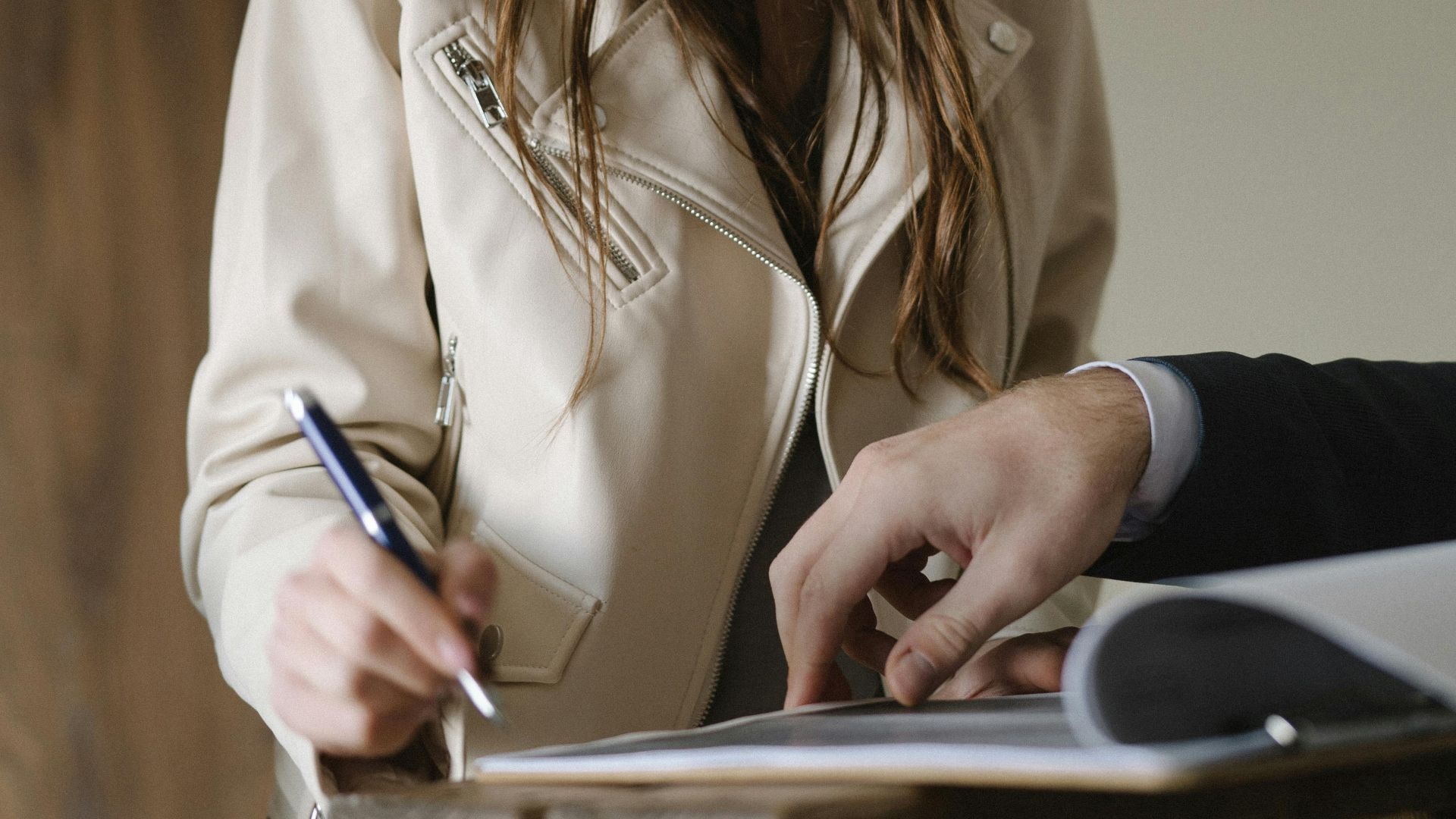 A young woman signing a business contract at an indoor table, holding a pen with focus and intent.