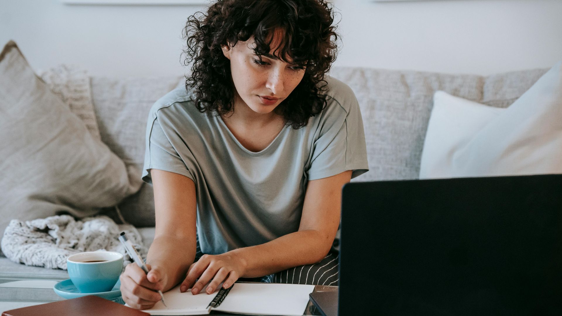 Focused young female student with dark curly hair in casual clothes taking notes in copybook while preparing for exams sitting on couch and using laptop at home